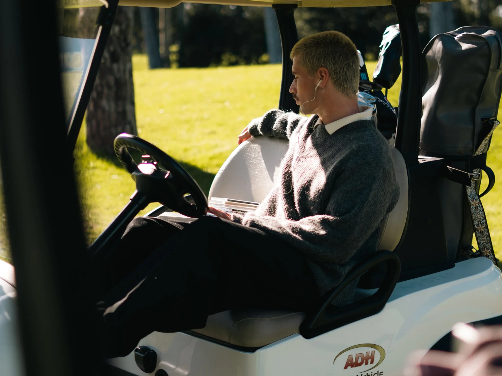 A man with short blonde hair wearing a gray sweater and white earbuds sitting on a golf cart with a black backpack attached to the back of the seat. The scenery outside is sunny with green grass and trees.
