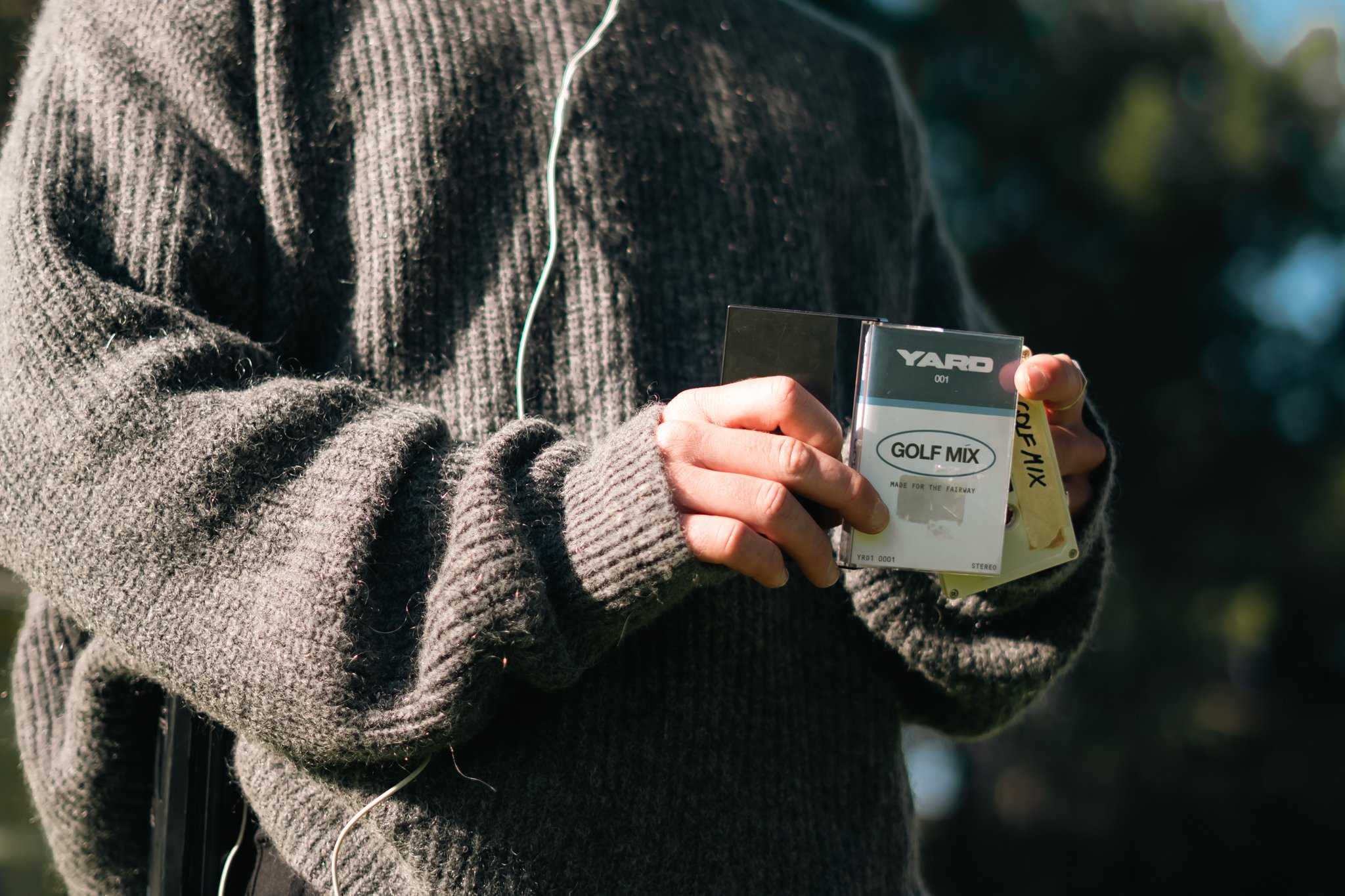Close-up of a person wearing a cozy, dark gray cable-knit sweater, holding a vintage compact case labeled 'YARD' with a pack of golf mix beside it, outdoors with blurred green trees in the background.