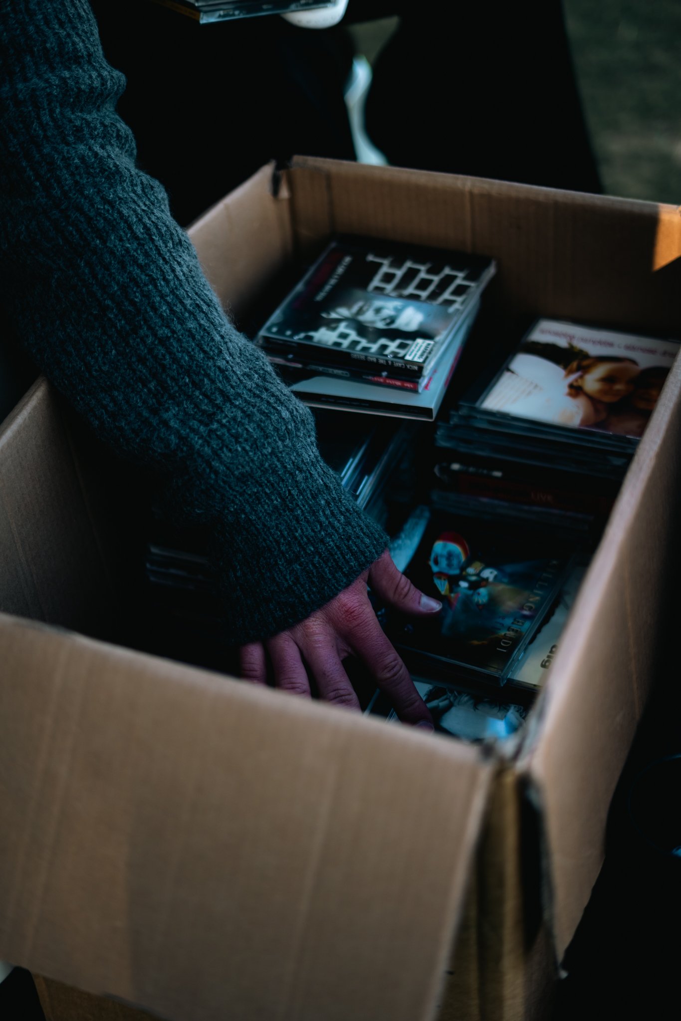 Close-up of a person's hand reaching into a cardboard box filled with stacked DVDs and Blu-ray discs, wearing a gray knitted sweater.