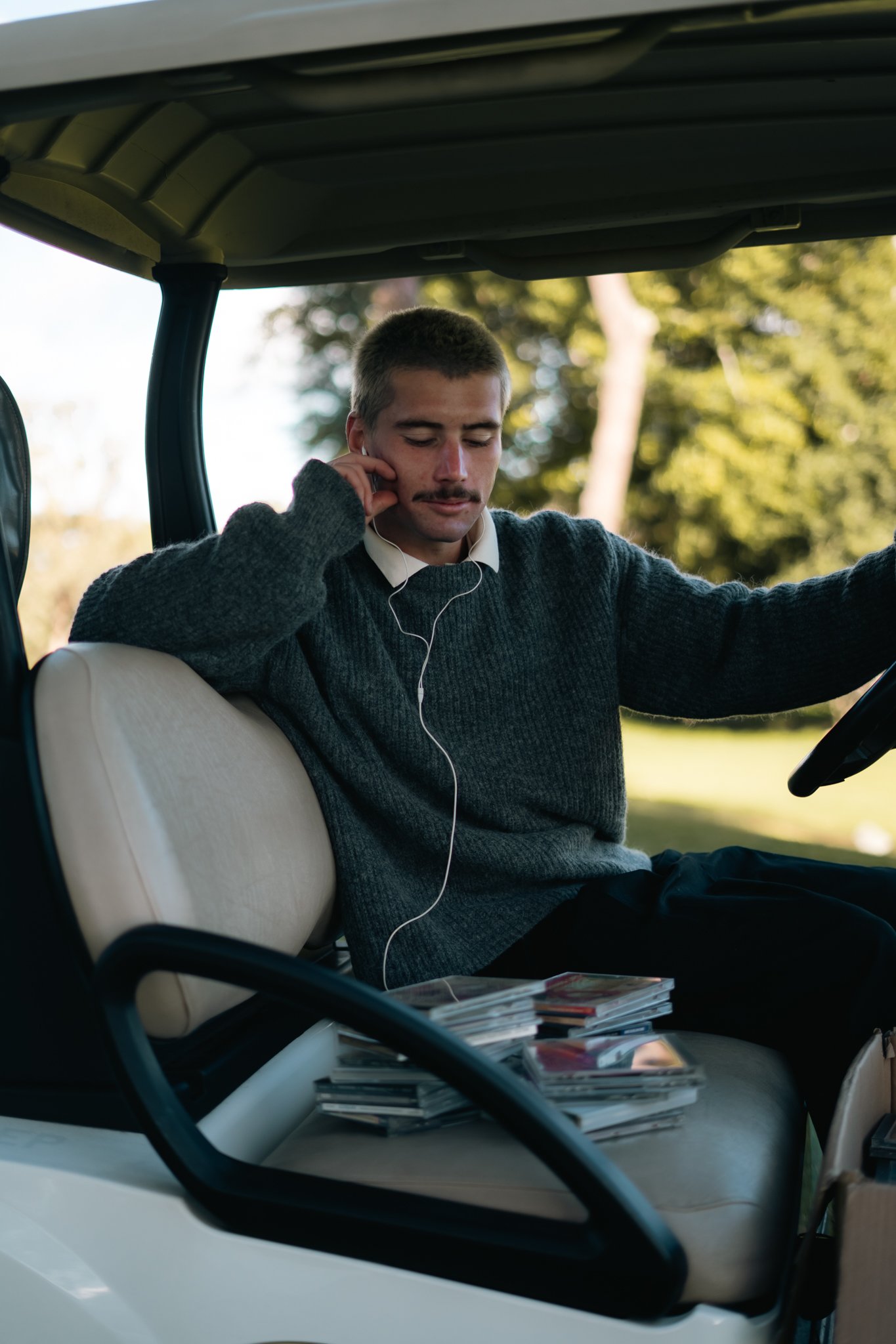 A man with a mustache and short hair sits in a car, wearing a gray sweater over a white shirt. He has a concerned facial expression, with one hand touching his face and earbud in his ear. The car's interior includes a stack of magazines on the dashboard, and the background outside the car shows trees and sunlight.