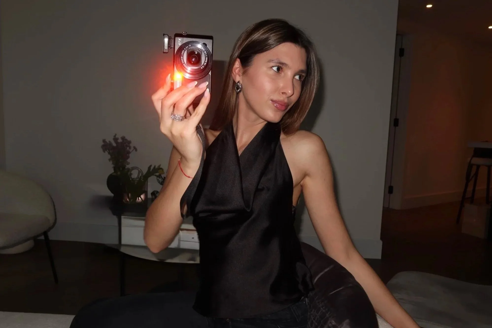 A woman in a black satin dress taking a selfie with a small vintage camera that has a red flash, sitting in a modern living room.