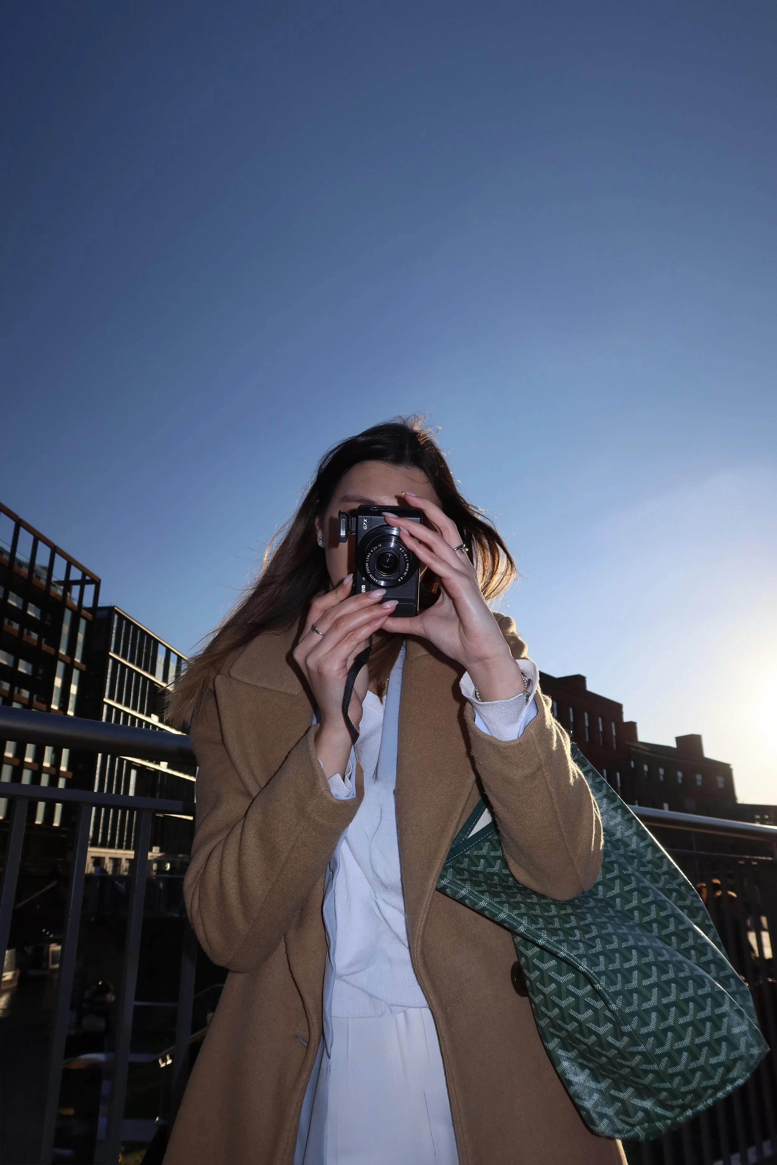 A woman taking a photo with a camera outdoors during daytime, wearing a tan coat, white shirt, and holding a patterned tote bag, with modern buildings and a clear blue sky in the background.