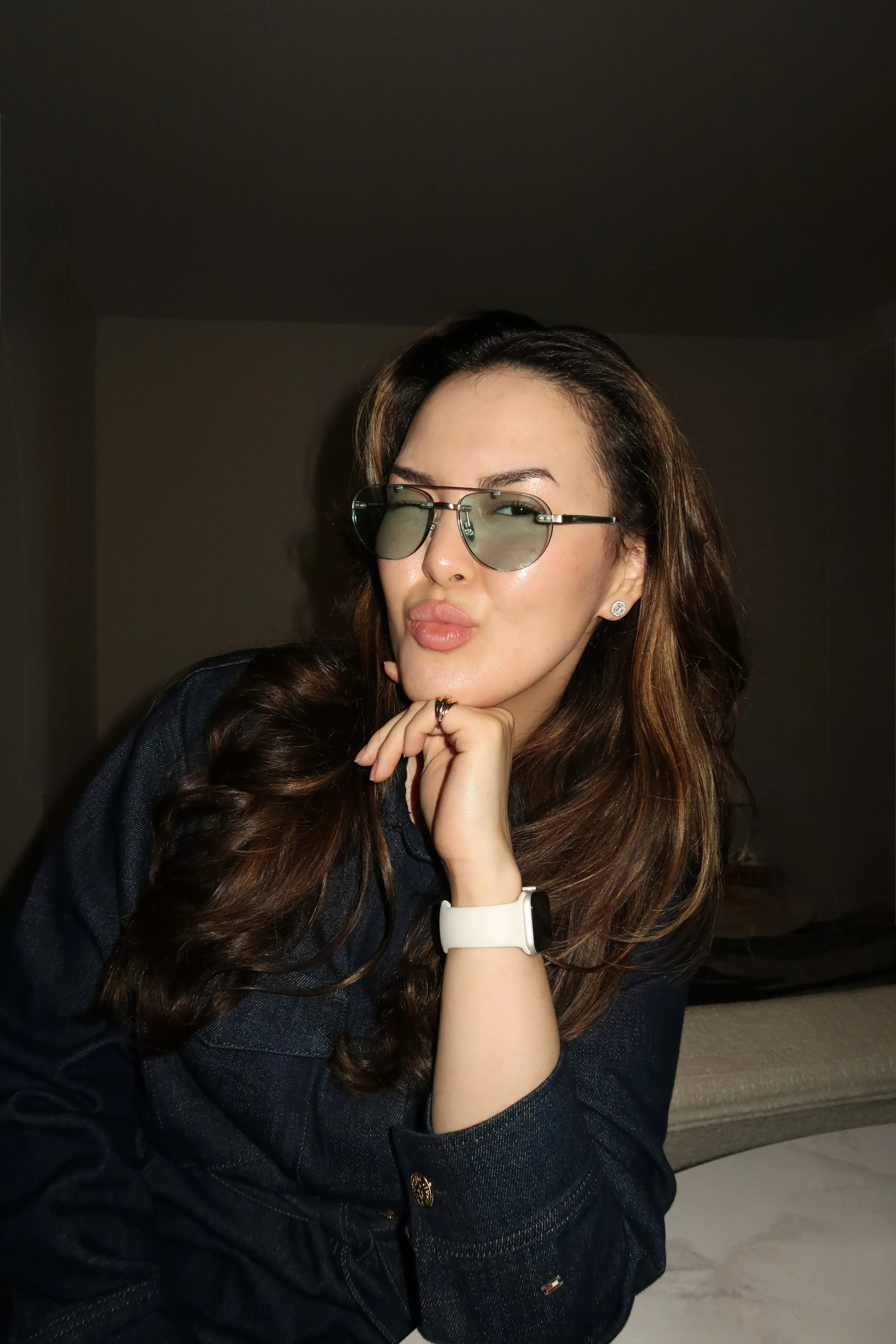 A young woman with long brown hair wearing round sunglasses, a dark denim jacket, a white watch, and earrings, sitting on a beige couch with her chin resting on her hand, indoors with a plain dark wall in the background.