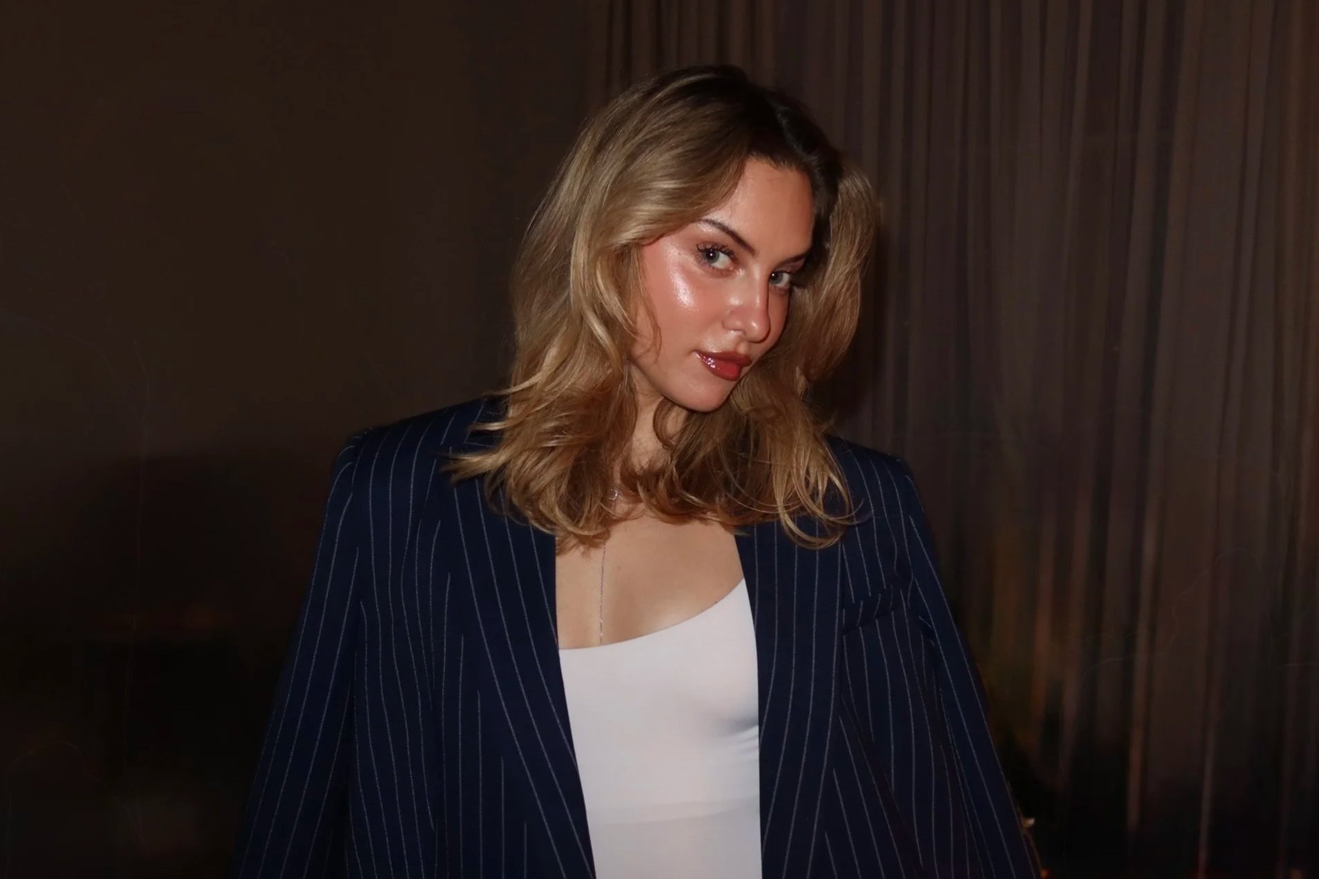 A woman with shoulder-length light brown hair and blue eyes, wearing makeup with nude lipstick, a white top, and a dark blue pinstripe blazer, standing in front of a wood-paneled background.