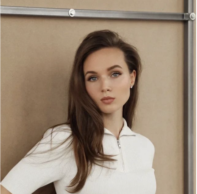 A young woman with long brown hair and blue eyes wearing a white zip-up top, standing against a plain beige background.