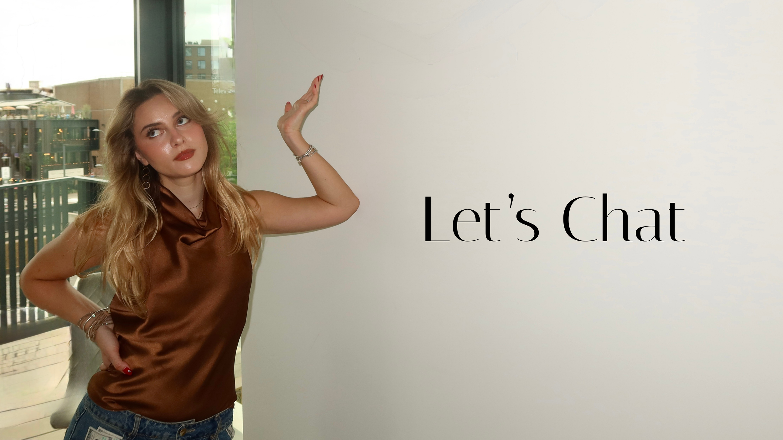 A woman with long blonde hair, red lipstick, and jewelry, wearing a satin brown top, stands next to a wall with the text "Let's Chat" in black font.