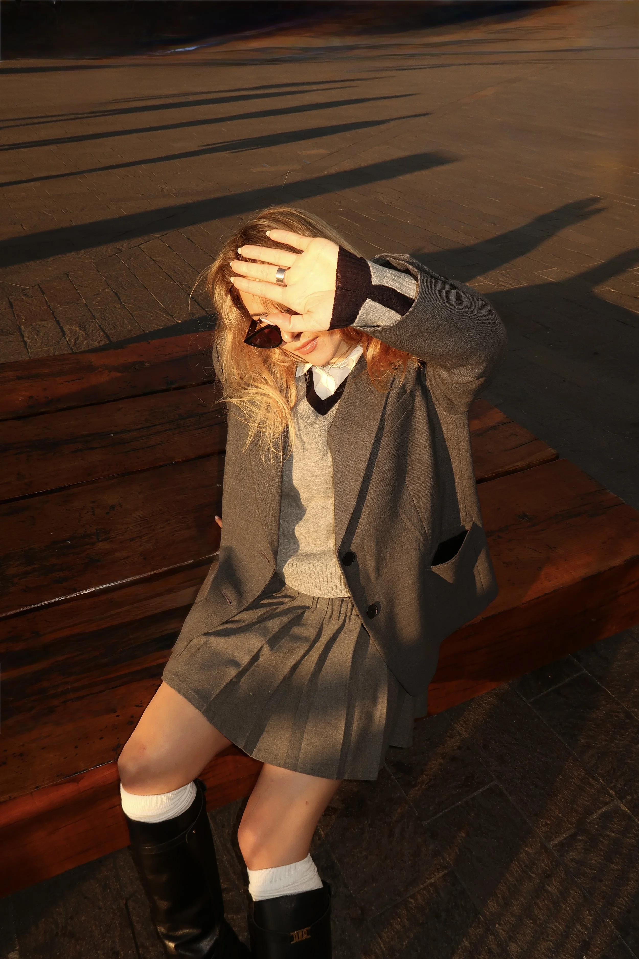 A woman sitting on a wooden bench outdoors, shielding her eyes from the sunlight with her hand. She is dressed in a gray blazer, matching skirt, white socks, and black boots, with sunglasses and wavy blonde hair. Shadows of the bench's slats are cast on the ground.
