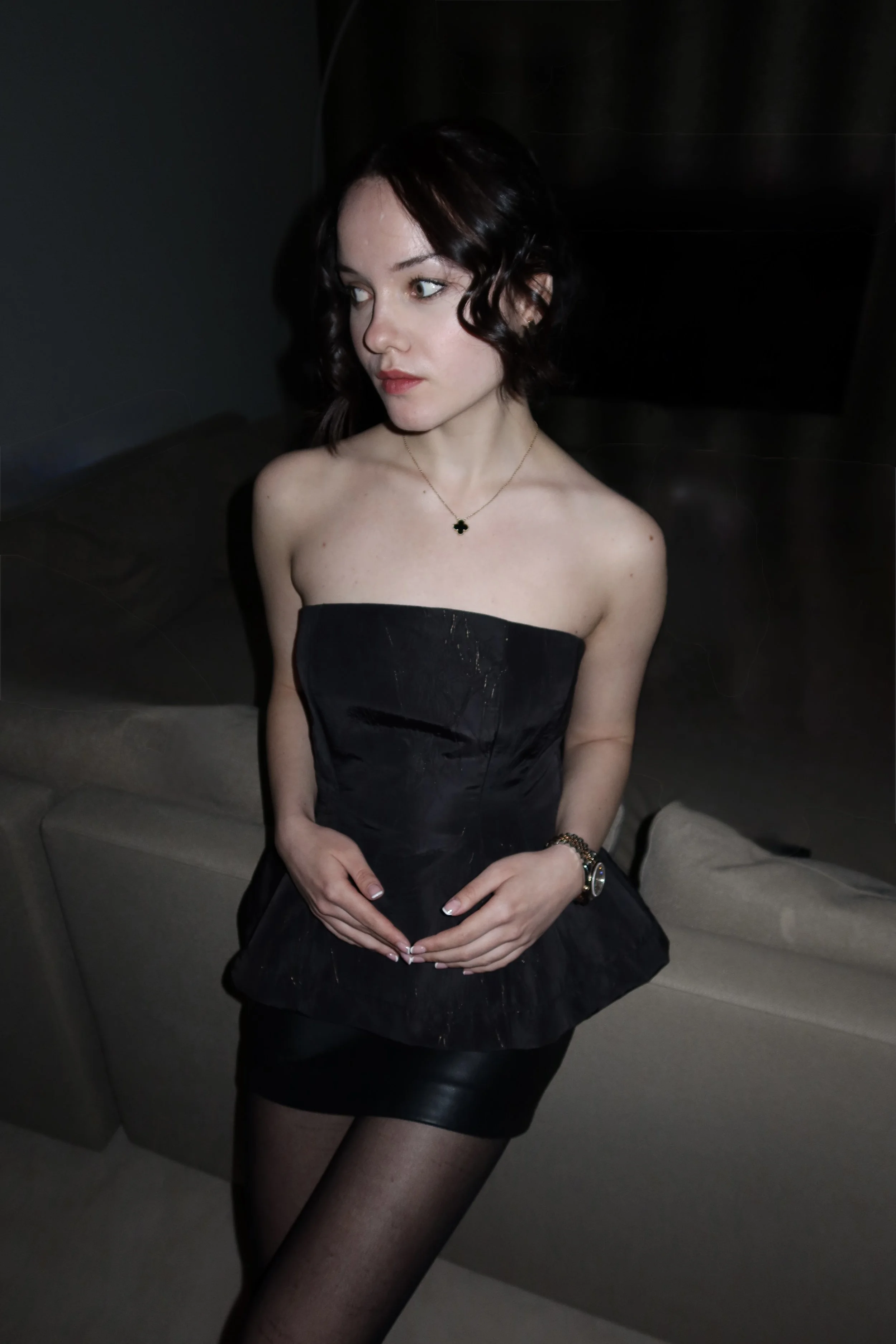 A woman with dark, curled hair wearing a black strapless dress, black stockings, and jewelry, standing in a dark room with a beige sofa in the background.