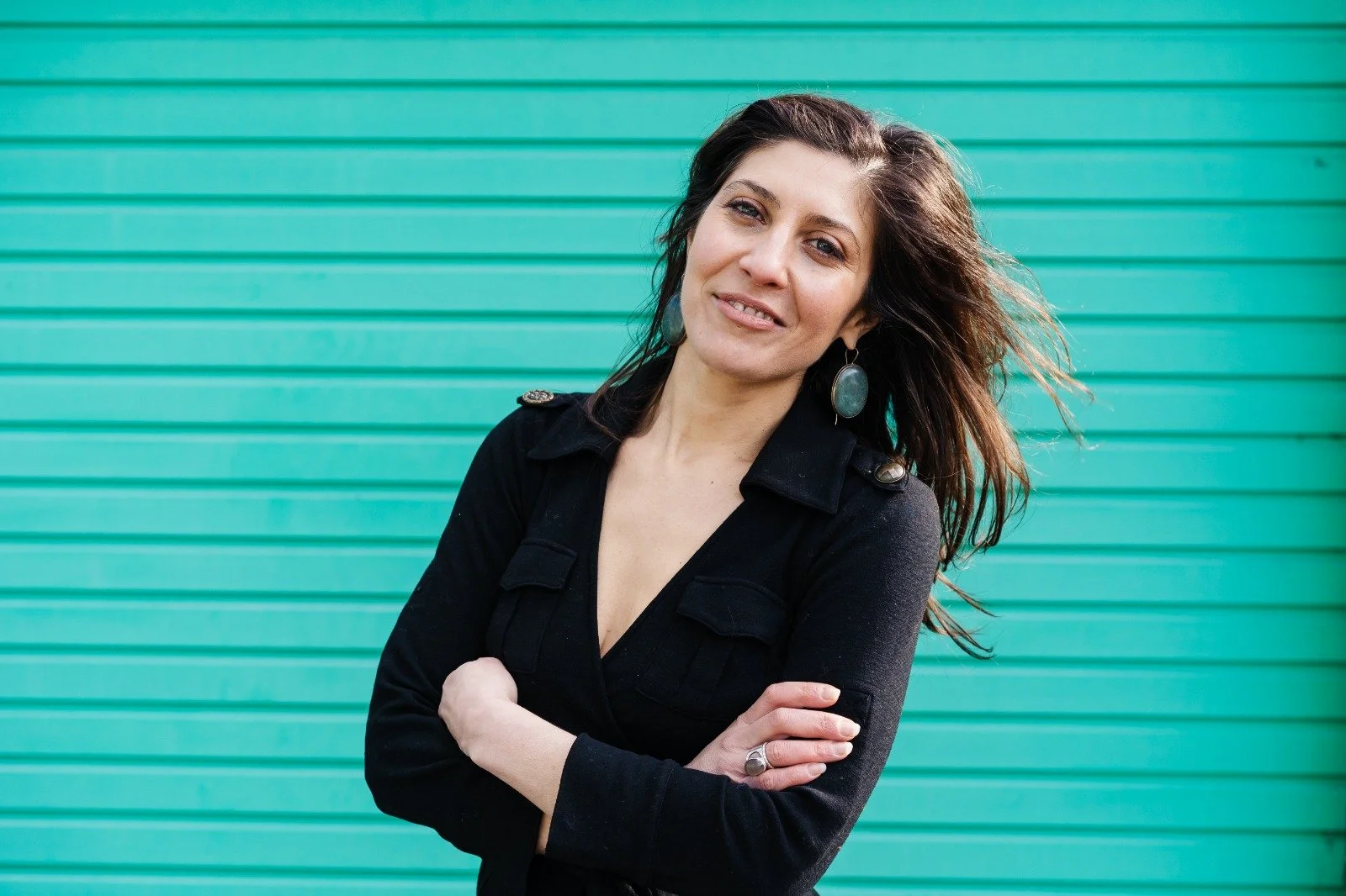 A smiling woman with dark hair, wearing a black jacket with epaulets and turquoise earrings, standing with arms crossed in front of a turquoise ribbed wall.