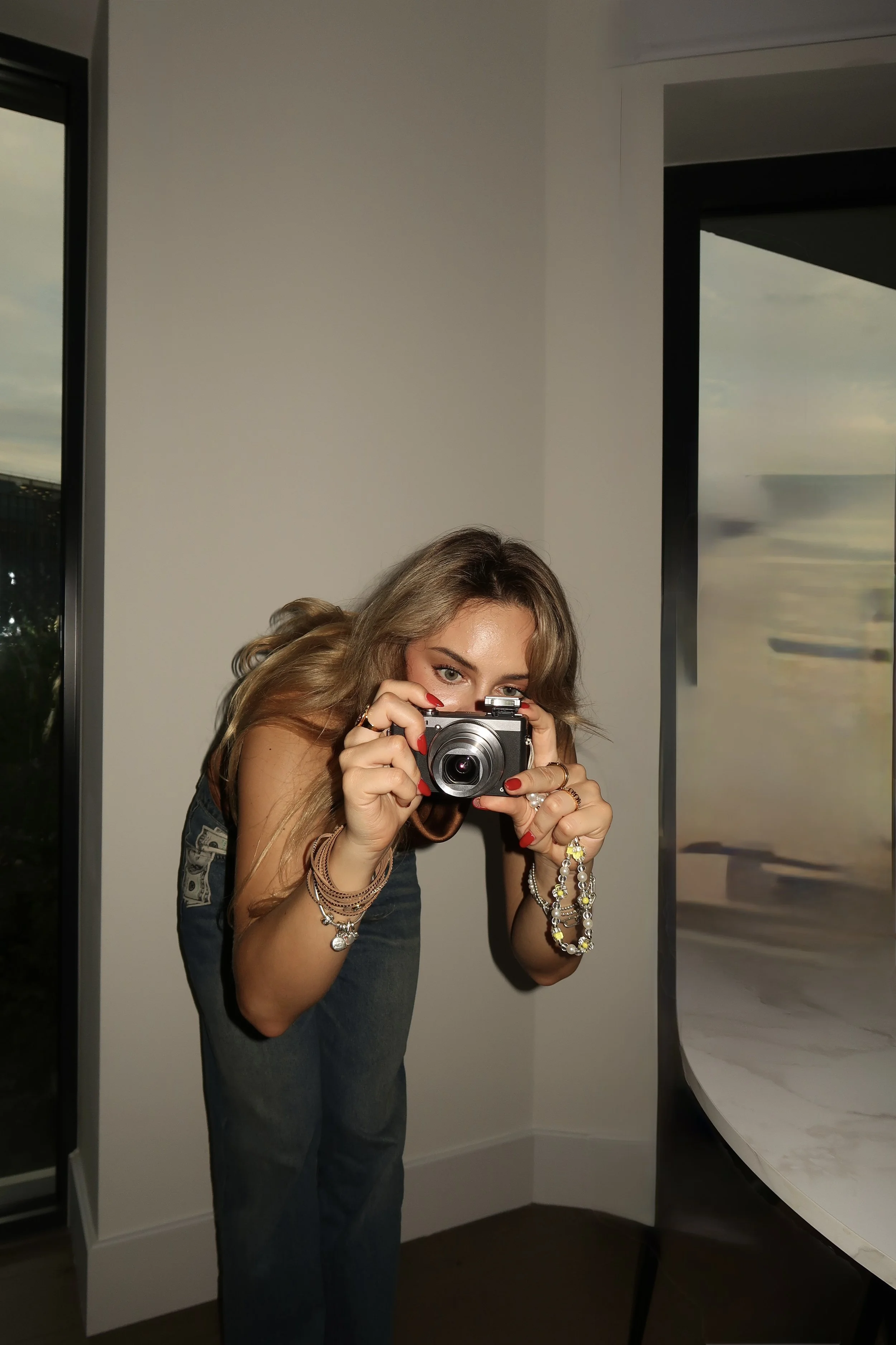 A woman with long wavy blonde hair taking a photo with a camera, wearing multiple bracelets and rings, standing near a window in a room with white walls and a curved white table.