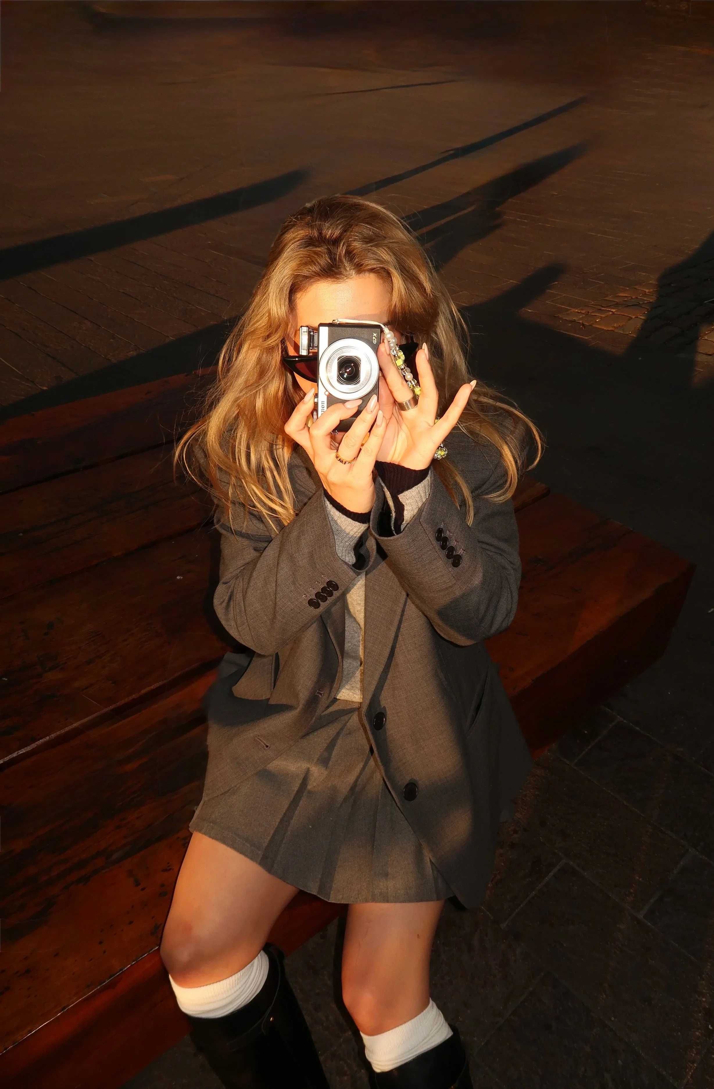 A young woman with long blonde hair, wearing a gray blazer and skirt with black shoes and white socks, is sitting on a wooden bench, taking a photo with a digital camera. The shot is taken outdoors with shadows cast on the ground.