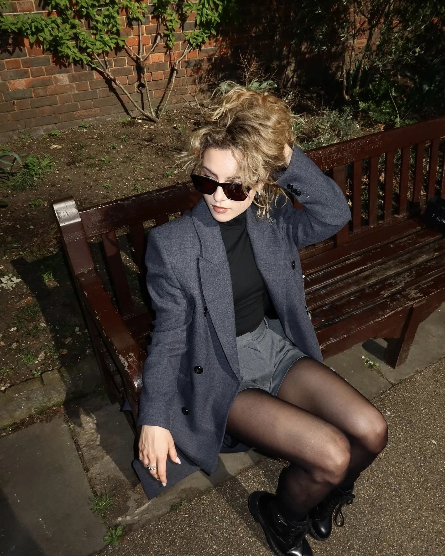 A woman sitting on a wooden park bench at night, wearing sunglasses, a grey blazer, black turtleneck, fishnet stockings, and black boots, with her hair styled in loose curls, outdoors near a brick wall and planted garden.