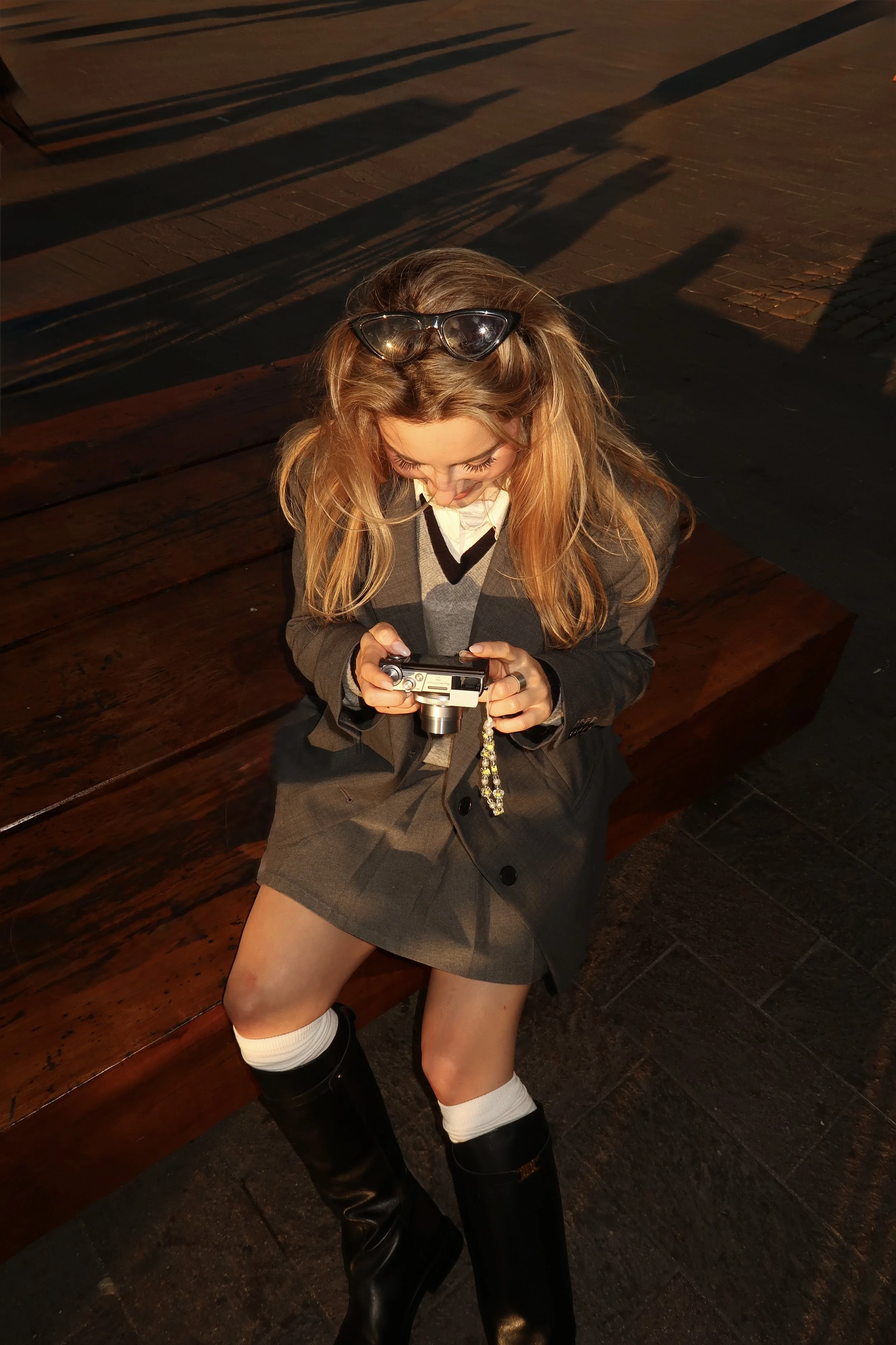 A woman sitting on a wooden bench wearing a gray plaid blazer, a matching skirt, white knee-high socks, and black boots, looking down at a camera she is holding.