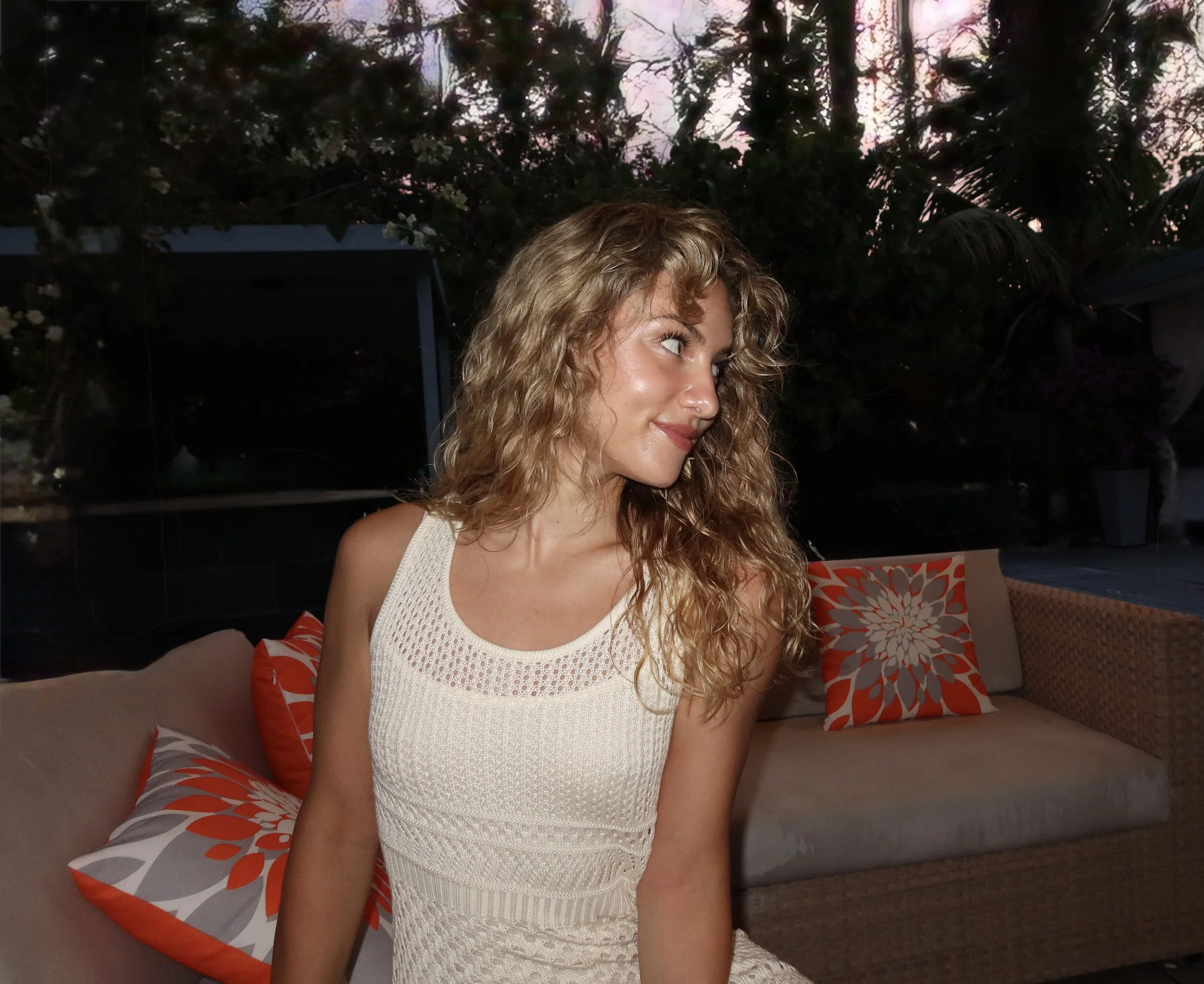 A woman with curly blonde hair sitting on a sofa with orange and gray patterned pillows, wearing a white knitted sleeveless top, with a dark outdoor scene visible in the background.