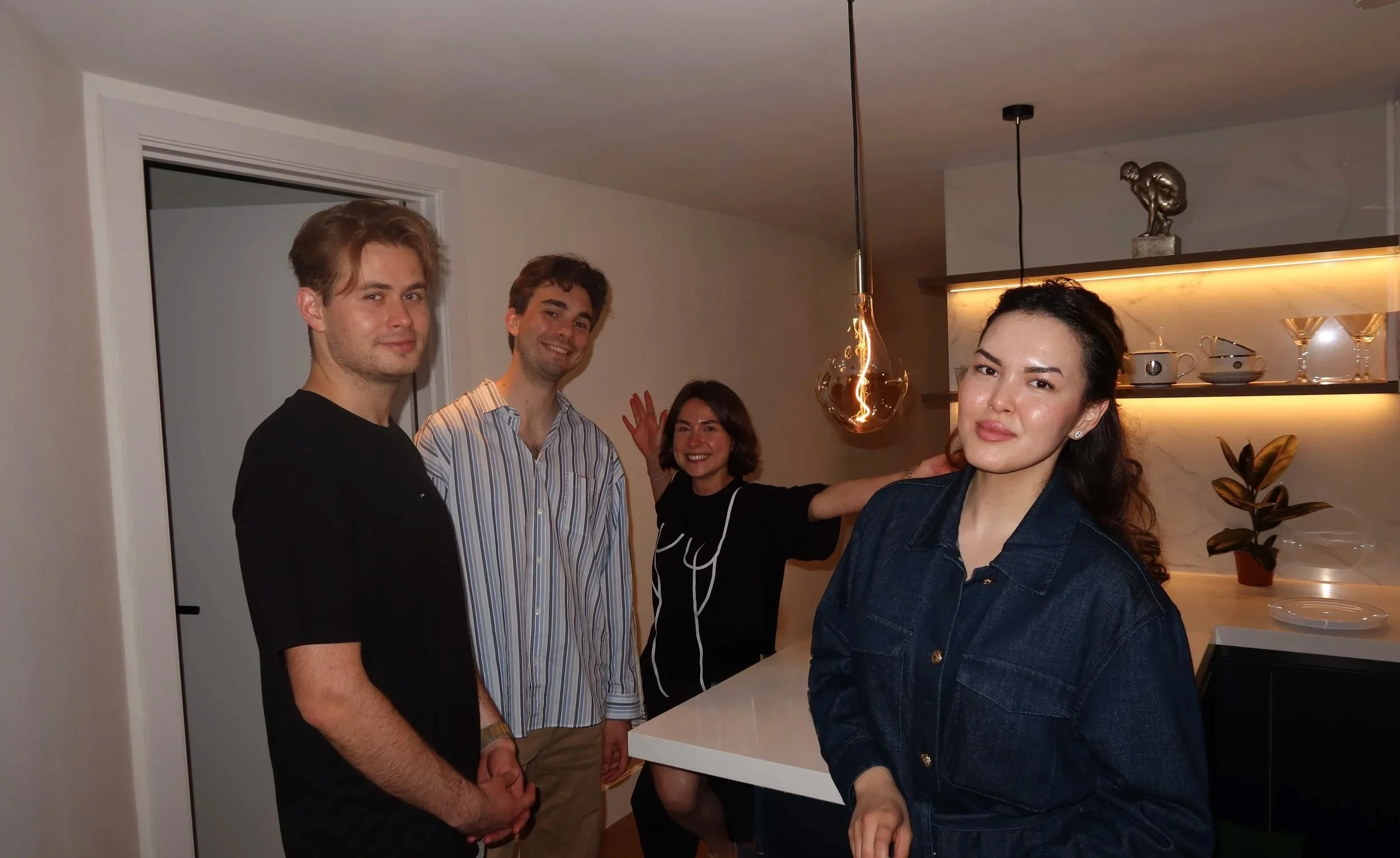 Four people gathered in a modern kitchen, smiling and posing for a photo. Two men are in the foreground, one in a black shirt and the other in a striped shirt. Two women are in the background, one waving and smiling, and another with dark hair and a dark jacket.