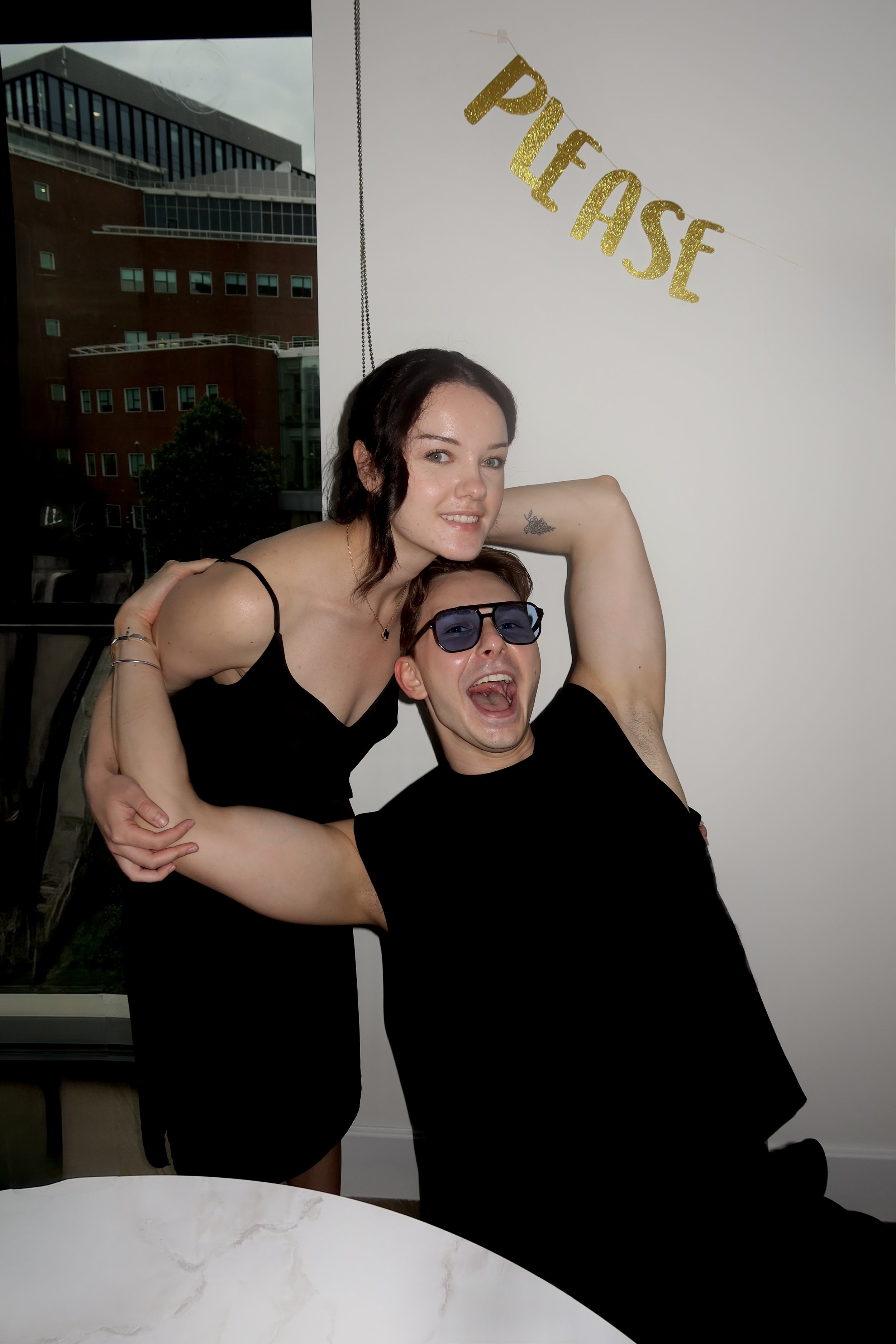A young woman with dark hair and a tattoo on her arm poses with a young man wearing sunglasses in front of a white wall with gold glittery "Party" banner. They are smiling and appear to be celebrating.