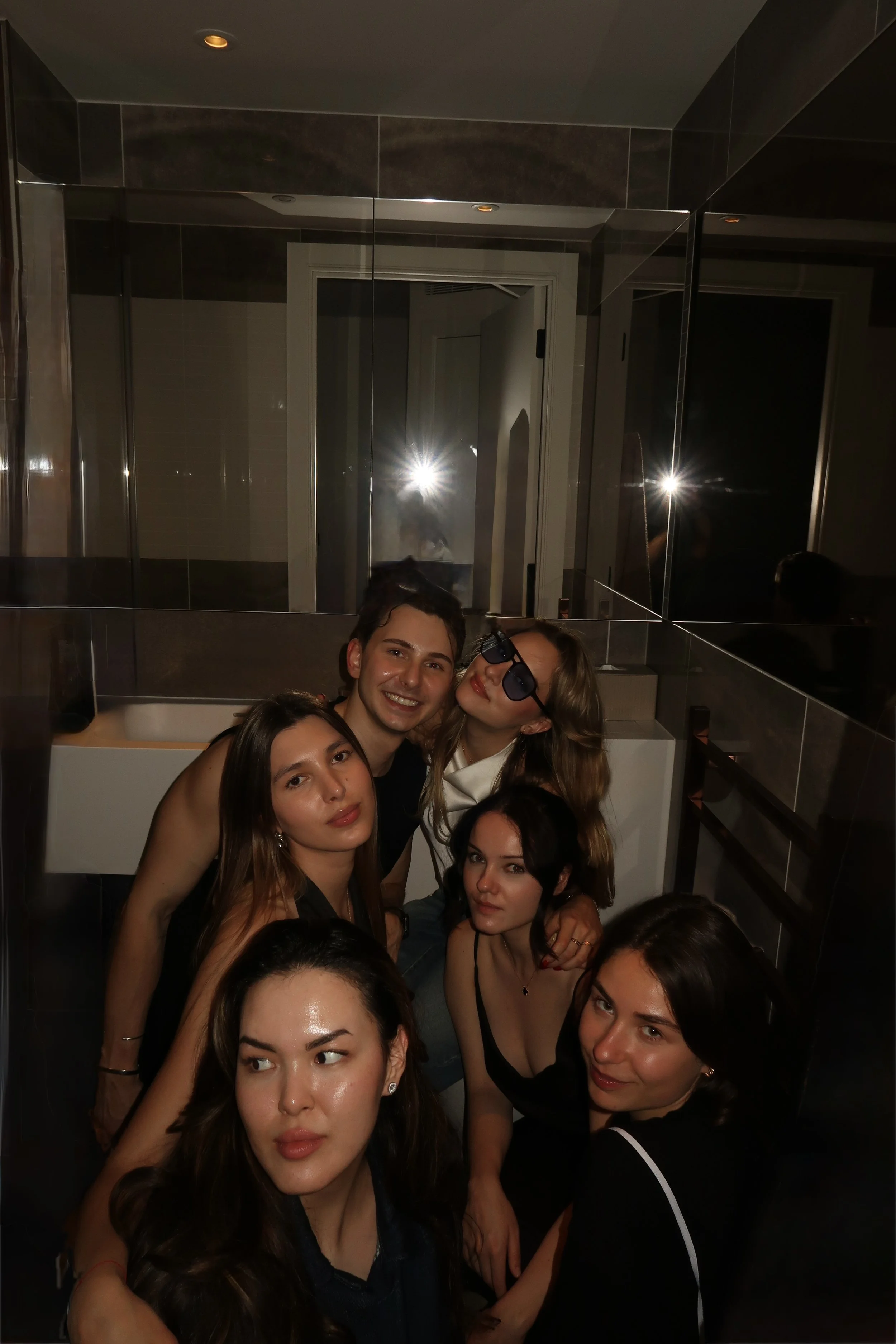 A group of young women and one young man posing for a photo in a dimly lit room with dark tiled walls and mirror panels, some making serious expressions and others smiling, with one woman in sunglasses. The photographer's flash is reflected in the mirror.