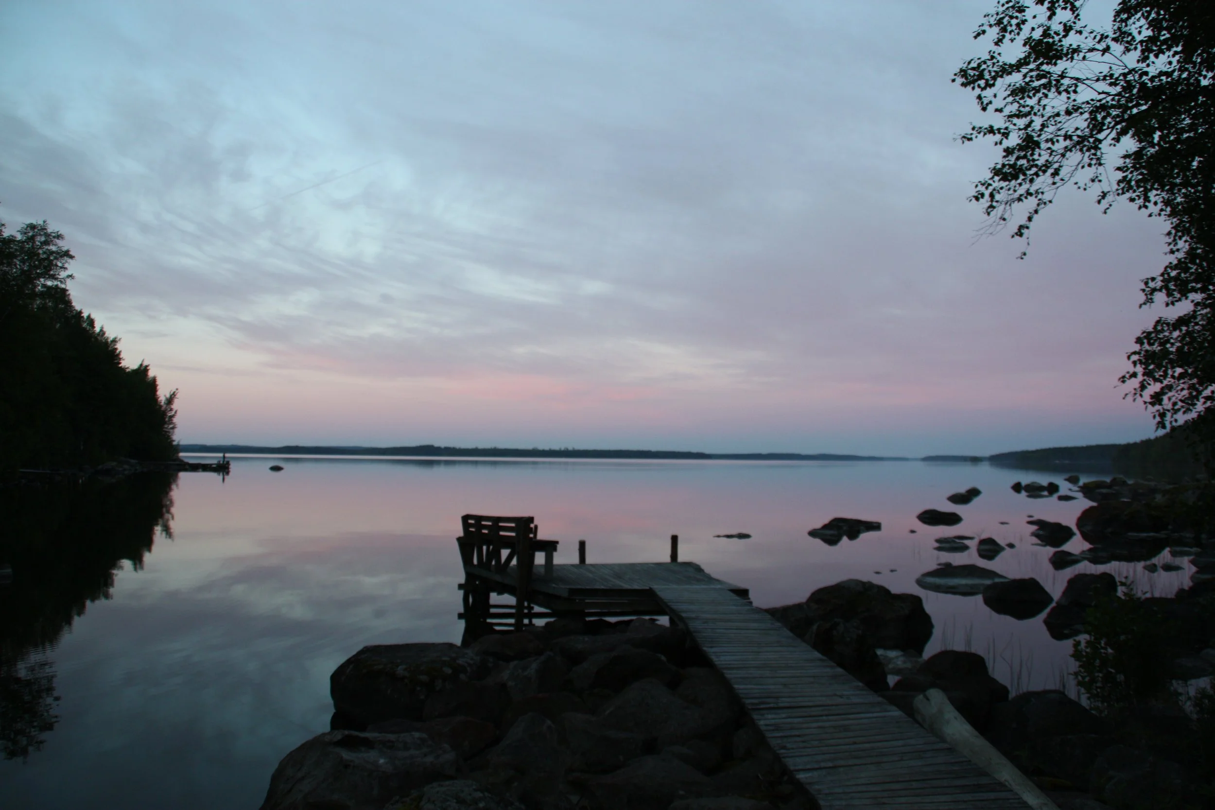 Abendstimmung an einem ruhigen See mit einem Steg und Bänken, umgeben von Bäumen, mit einem bewölkten Himmel in sanften Pastellfarben.