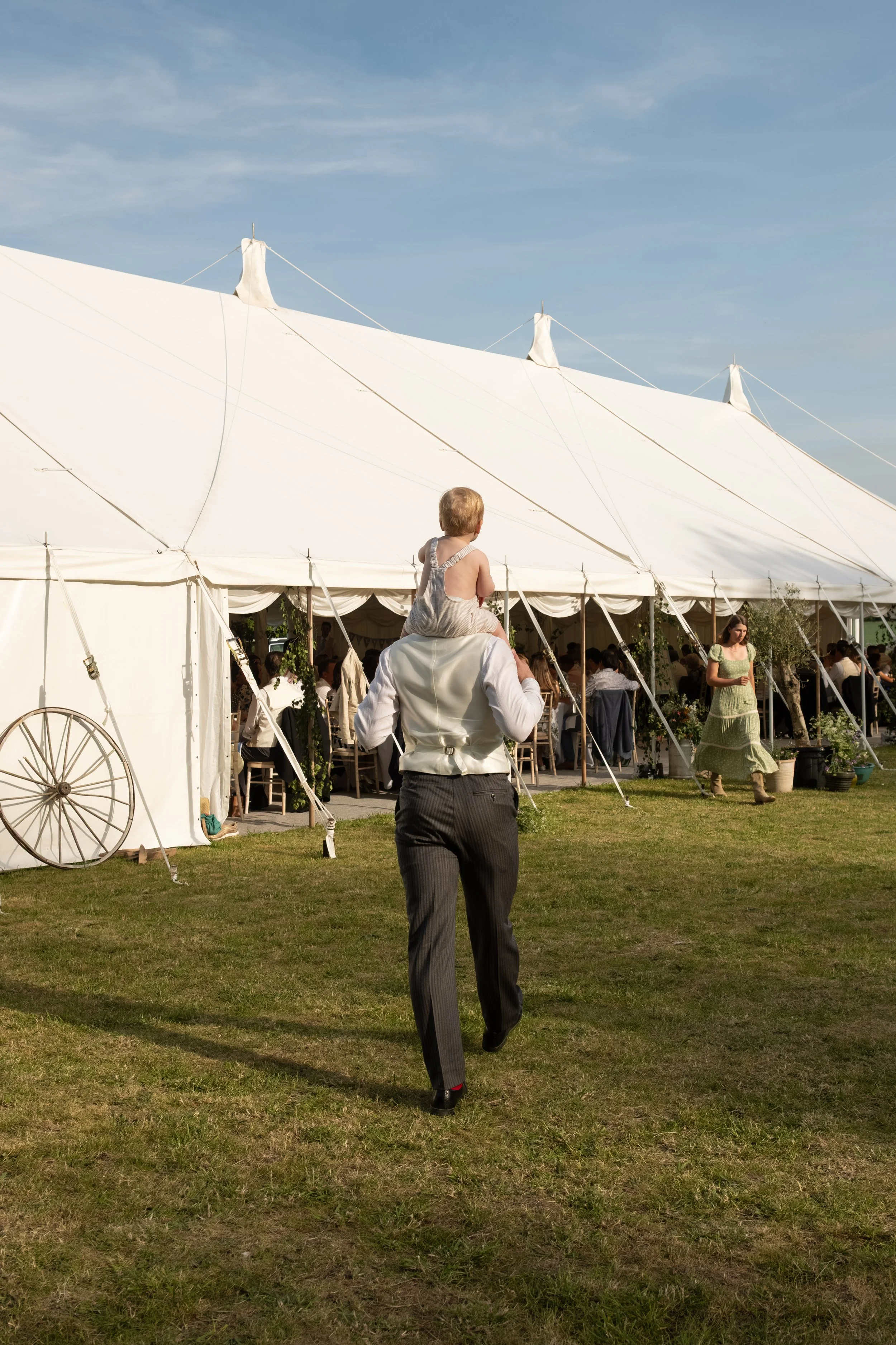 A man with a young girl sitting on his shoulders walking towards a white tent at an outdoor event on a sunny day, with clear skies and green grass.