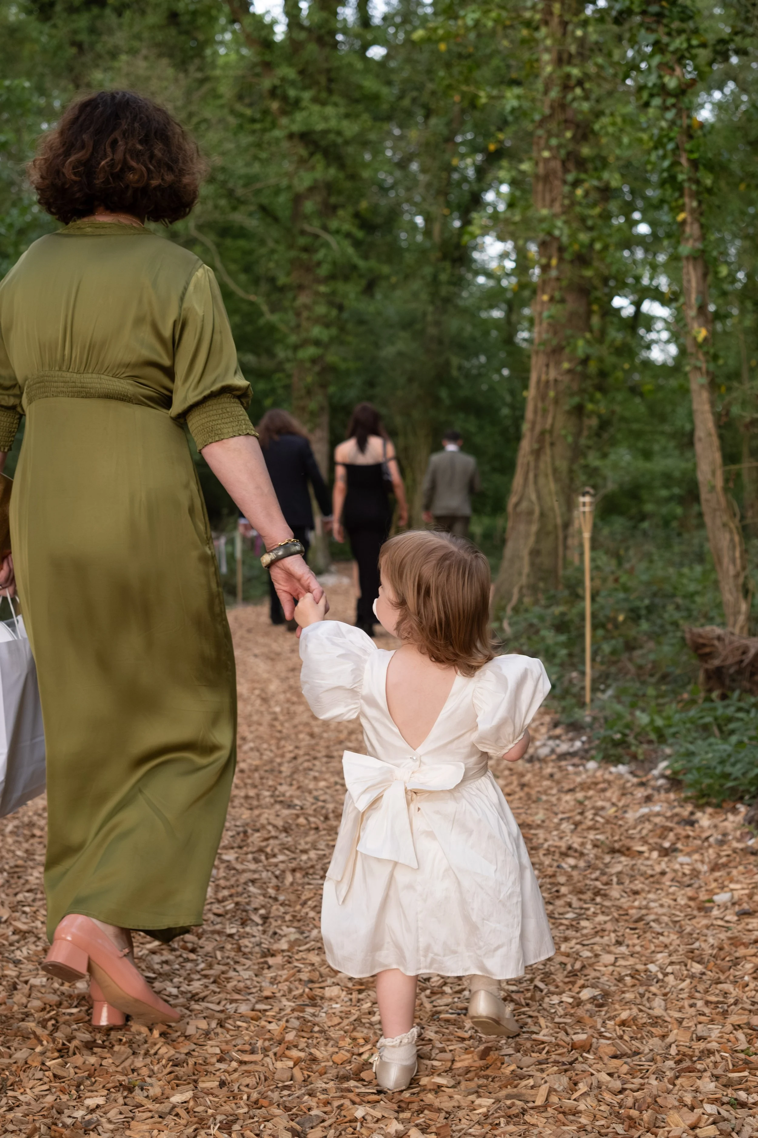 A woman in a green dress holding the hand of a young girl in a white dress walking on a leaf-covered path in a wooded area, with several other people in the background.