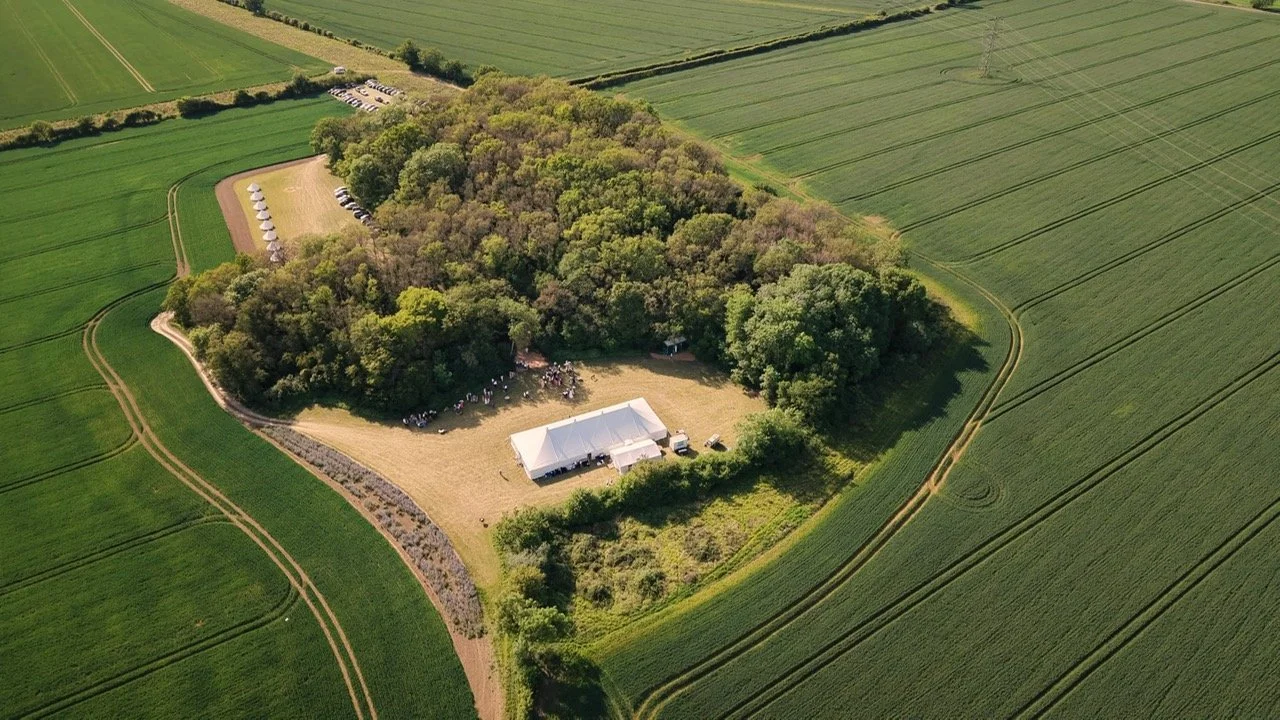 Aerial view of a large white event tent set up on a grassy patch surrounded by trees, with a parking lot nearby and expansive green farmland in the background.