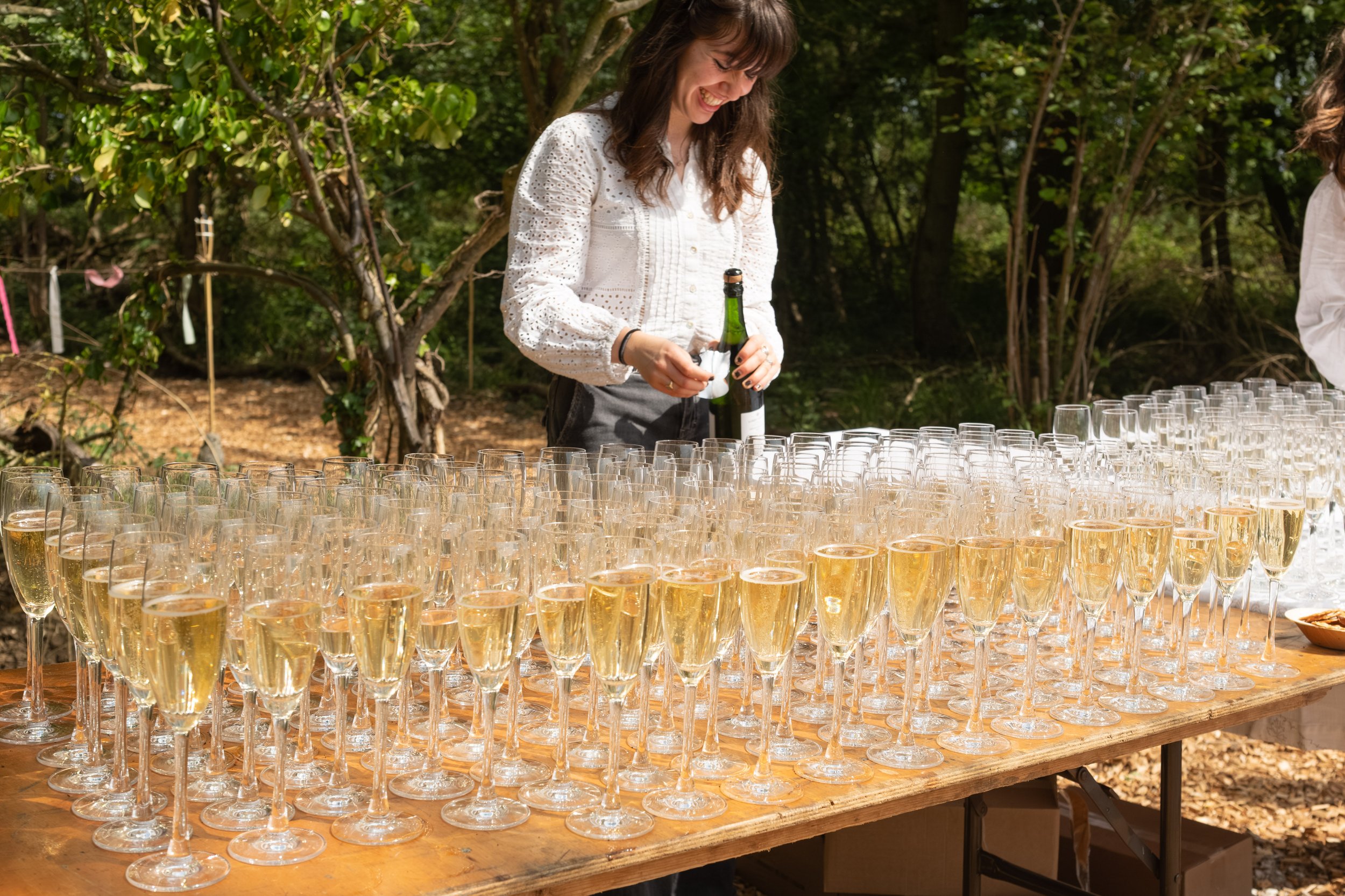 A woman smiling and holding a bottle of champagne at an outdoor celebration with many champagne glasses on a wooden table.