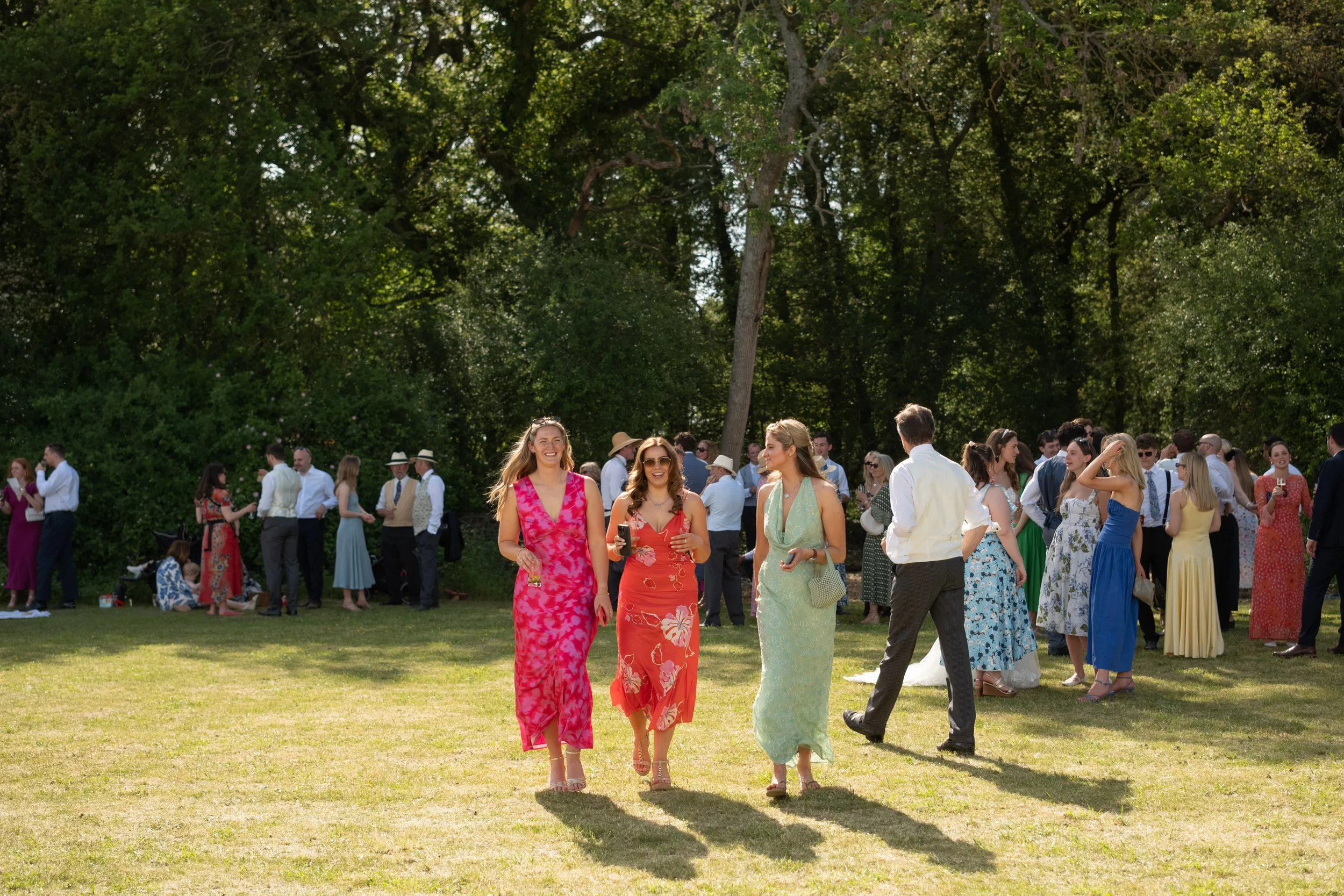 Several women in colorful dresses walking on a grassy lawn at an outdoor gathering, with many people mingling and chatting in the background under tall, leafy trees.