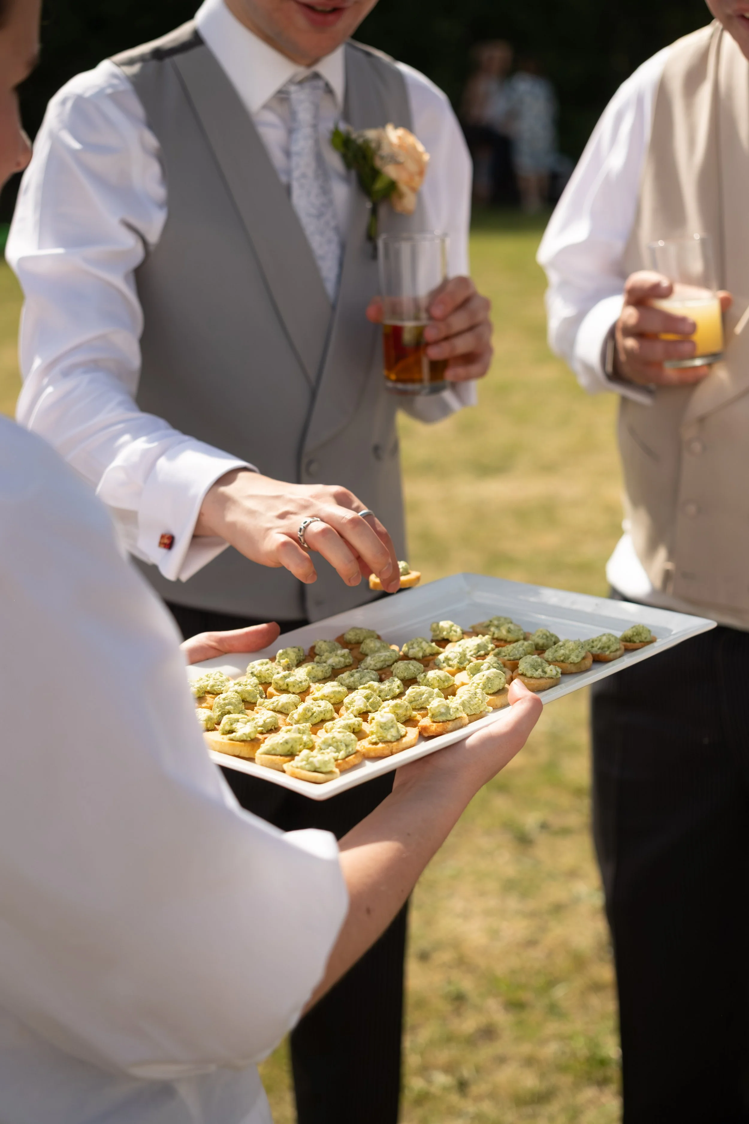 People dressed in formal wedding attire sharing drinks and serving appetizers outdoors at a garden wedding reception.
