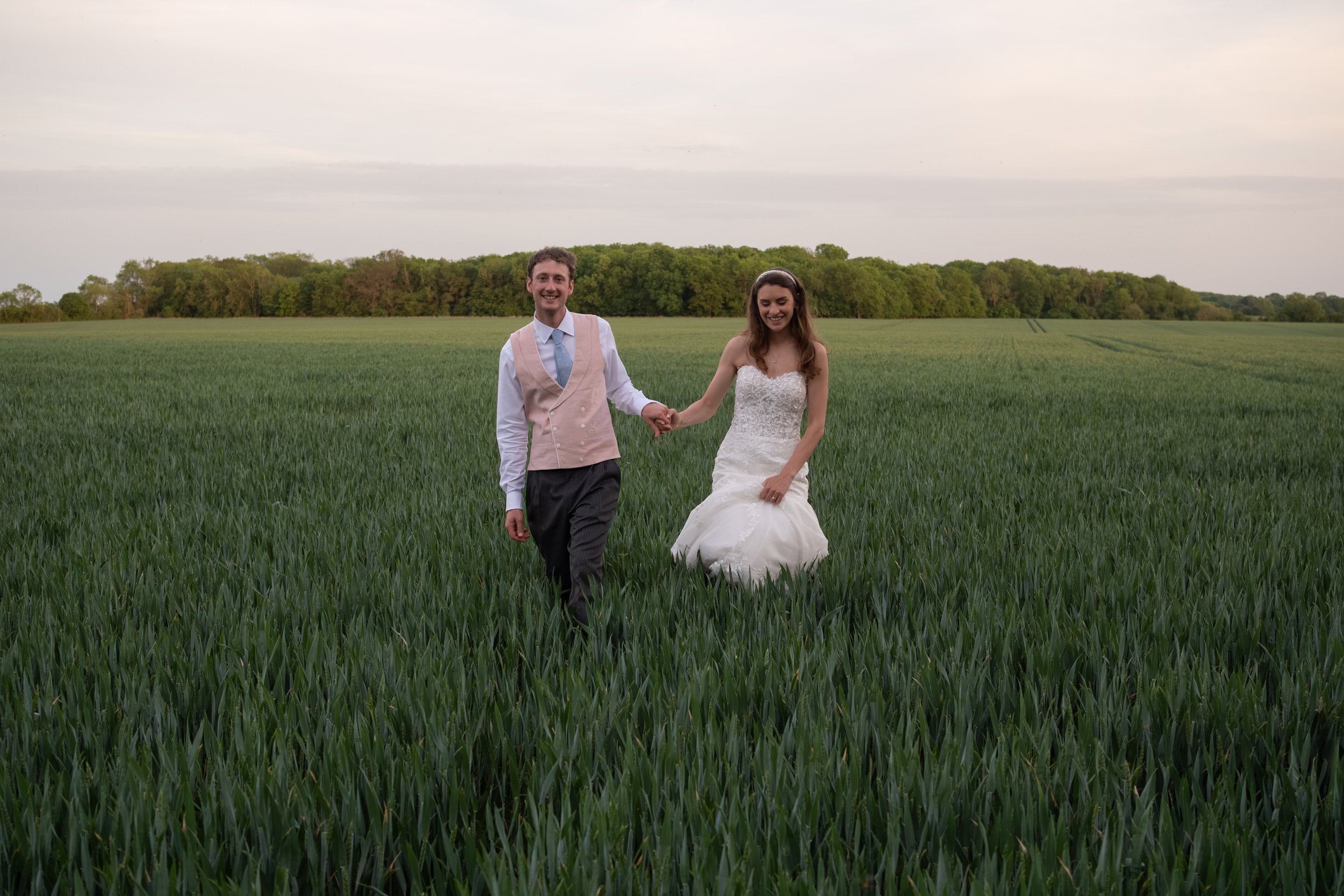 A smiling man and woman holding hands in a green field. The woman is wearing a white wedding dress, and the man is wearing a pink vest with a blue tie and dark pants. There are trees in the background and an overcast sky.