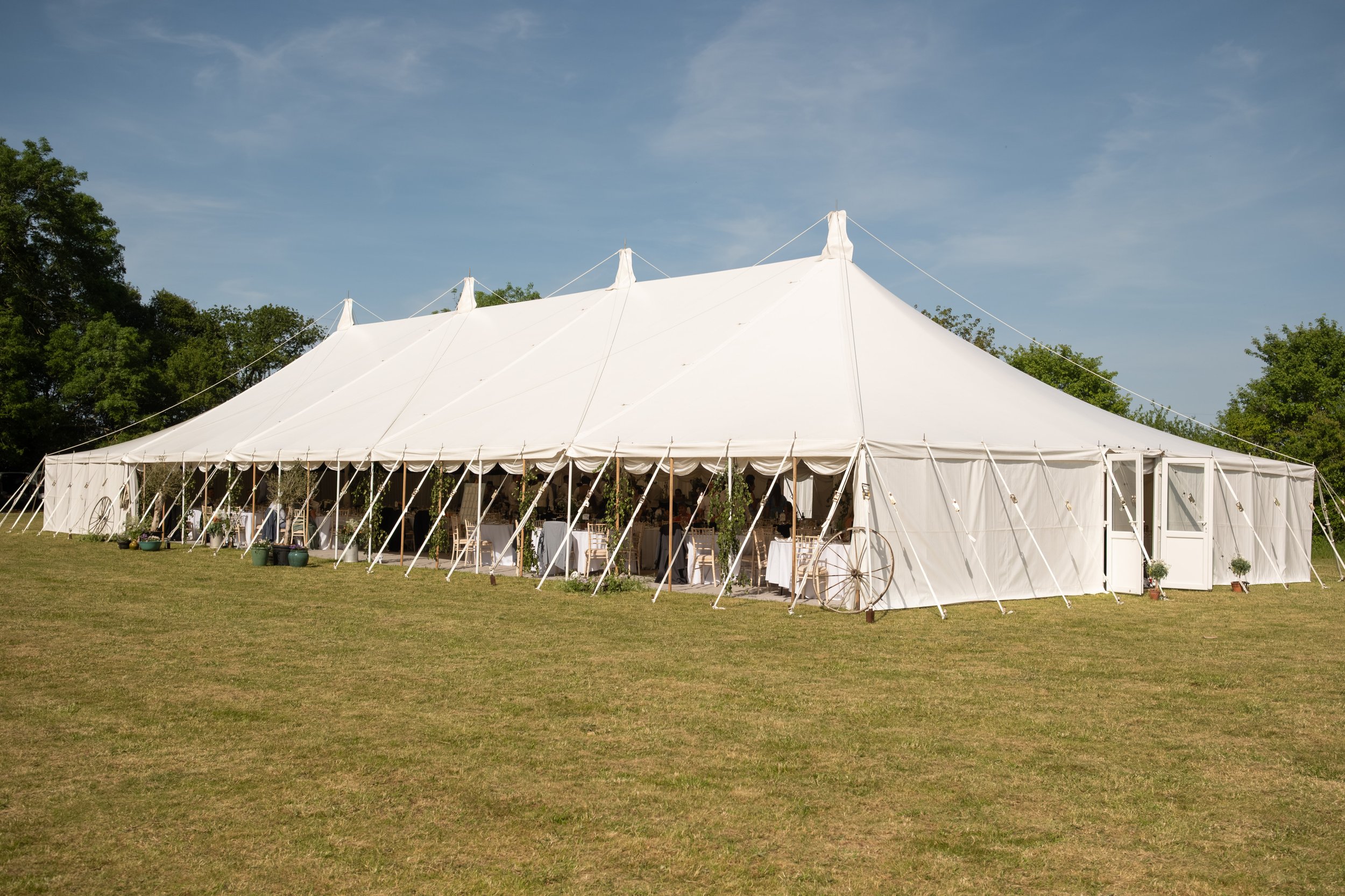 A large white event tent with open sides set up on a grassy field, surrounded by trees, under a partly cloudy sky.