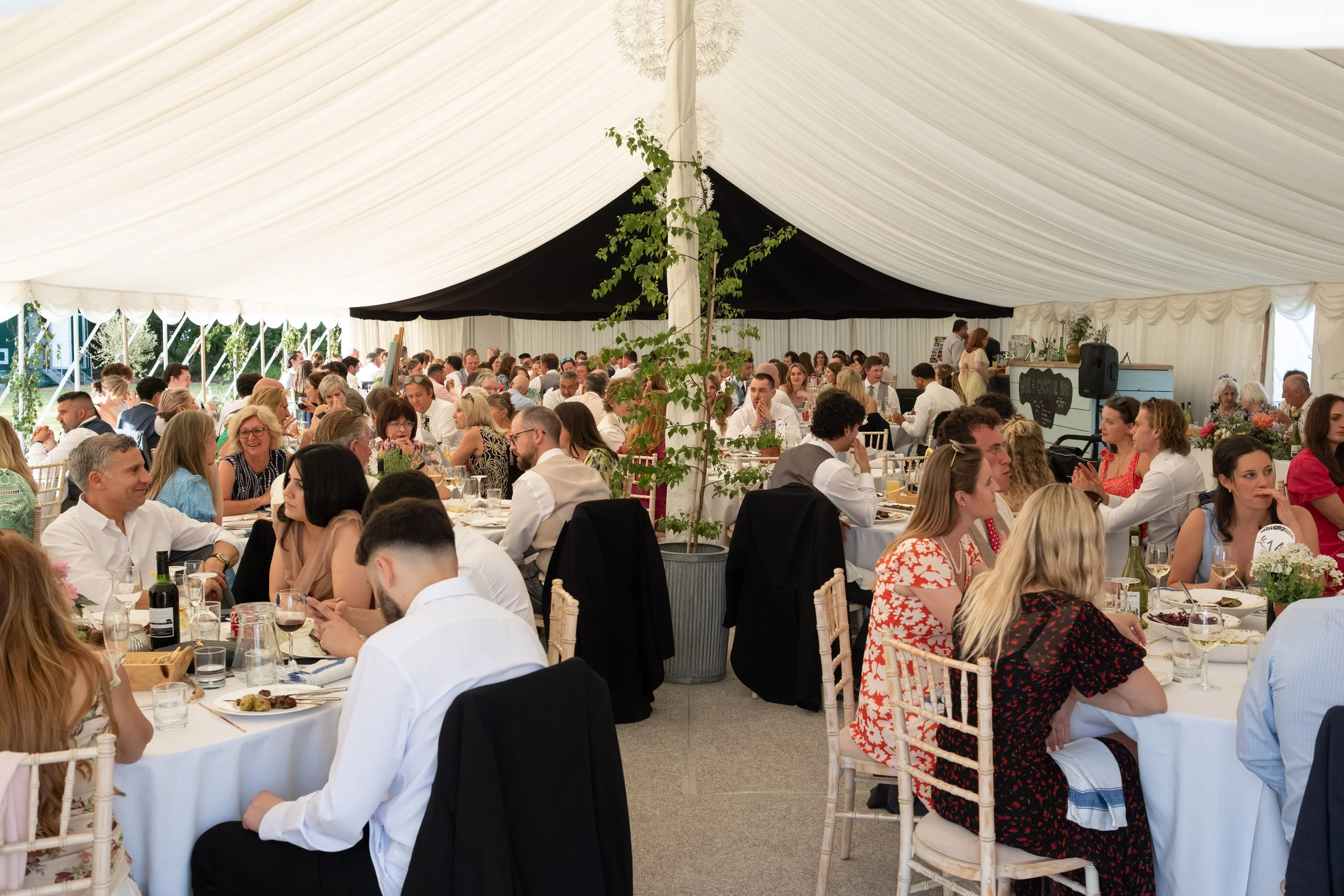 People sitting at tables inside a large white event tent during a celebration, with greenery outside visible through the tent sides.
