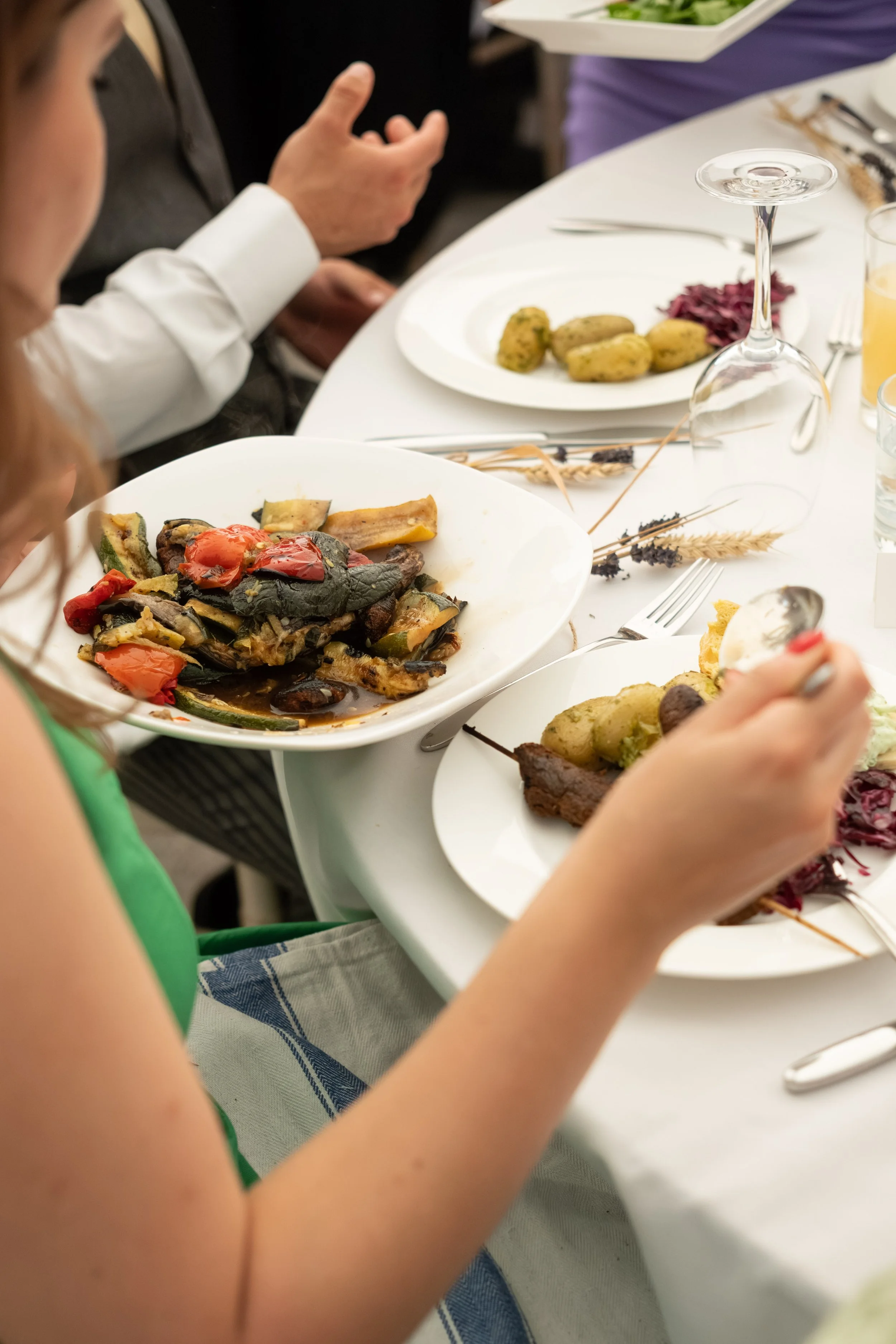 Close-up of a person in a white shirt at a dining table with dishes of cooked vegetables, potatoes, purple cabbage, and grilled meat, along with wine glasses and silverware.