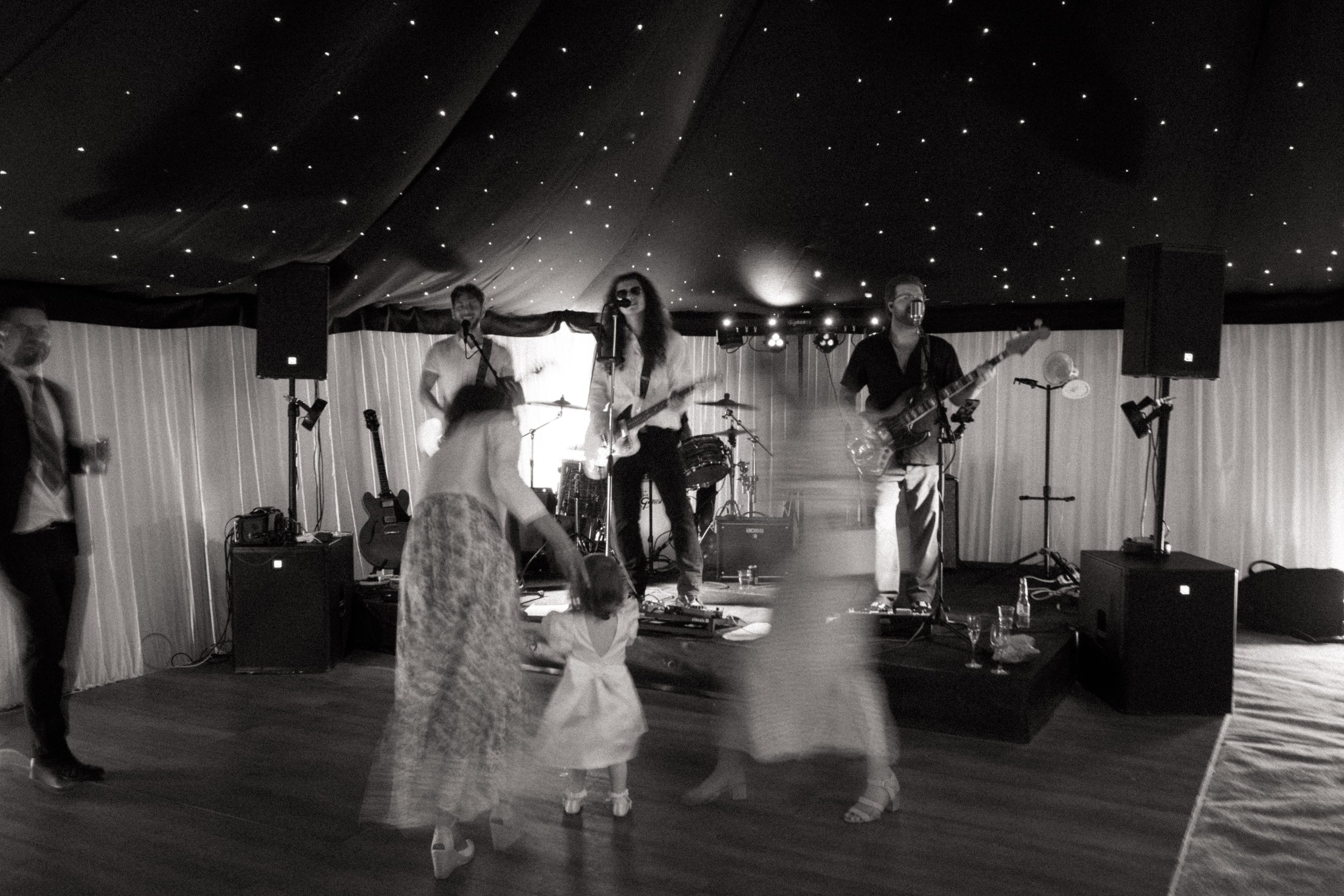 A live band performs on stage under a ceiling decorated with star-like lights, with performers playing instruments and a person dancing in front of the stage, in a dimly lit venue.