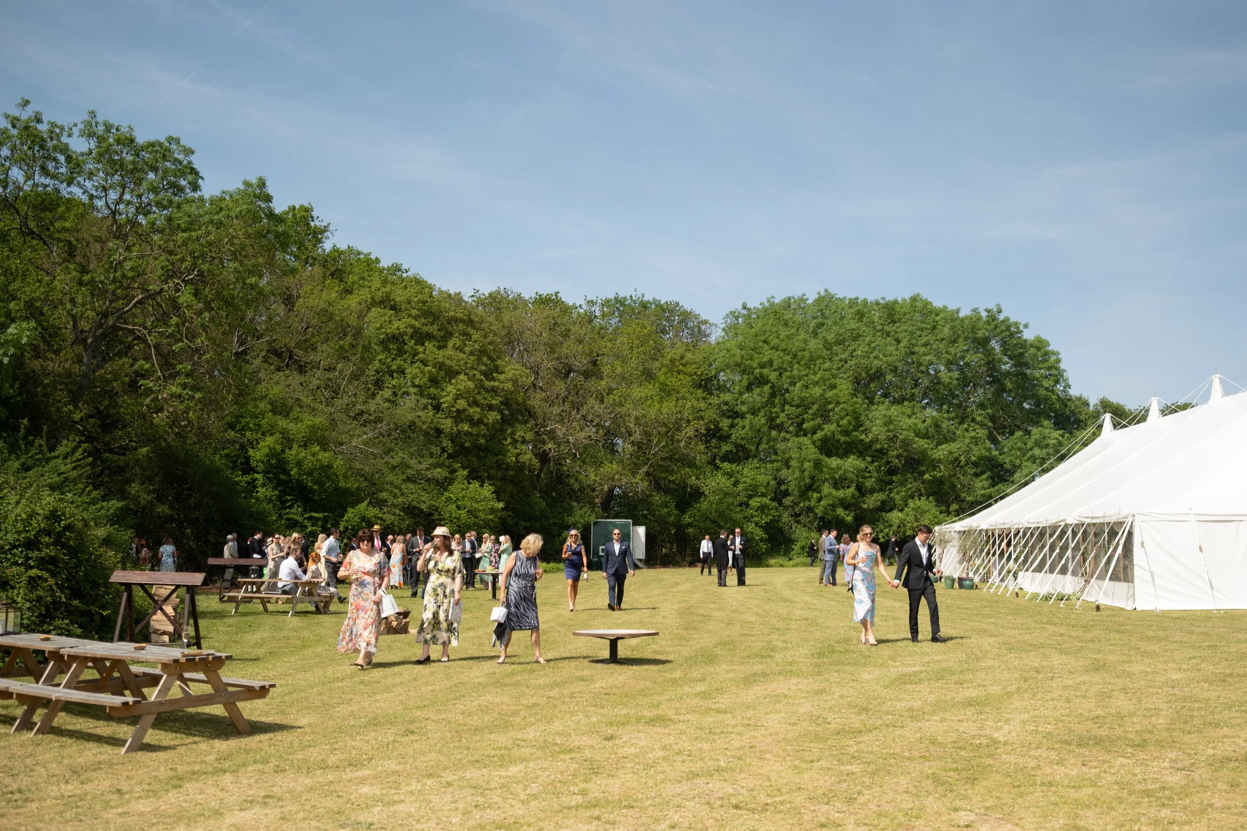 People attending an outdoor event near a large white tent on a grassy field, with trees and a blue sky.