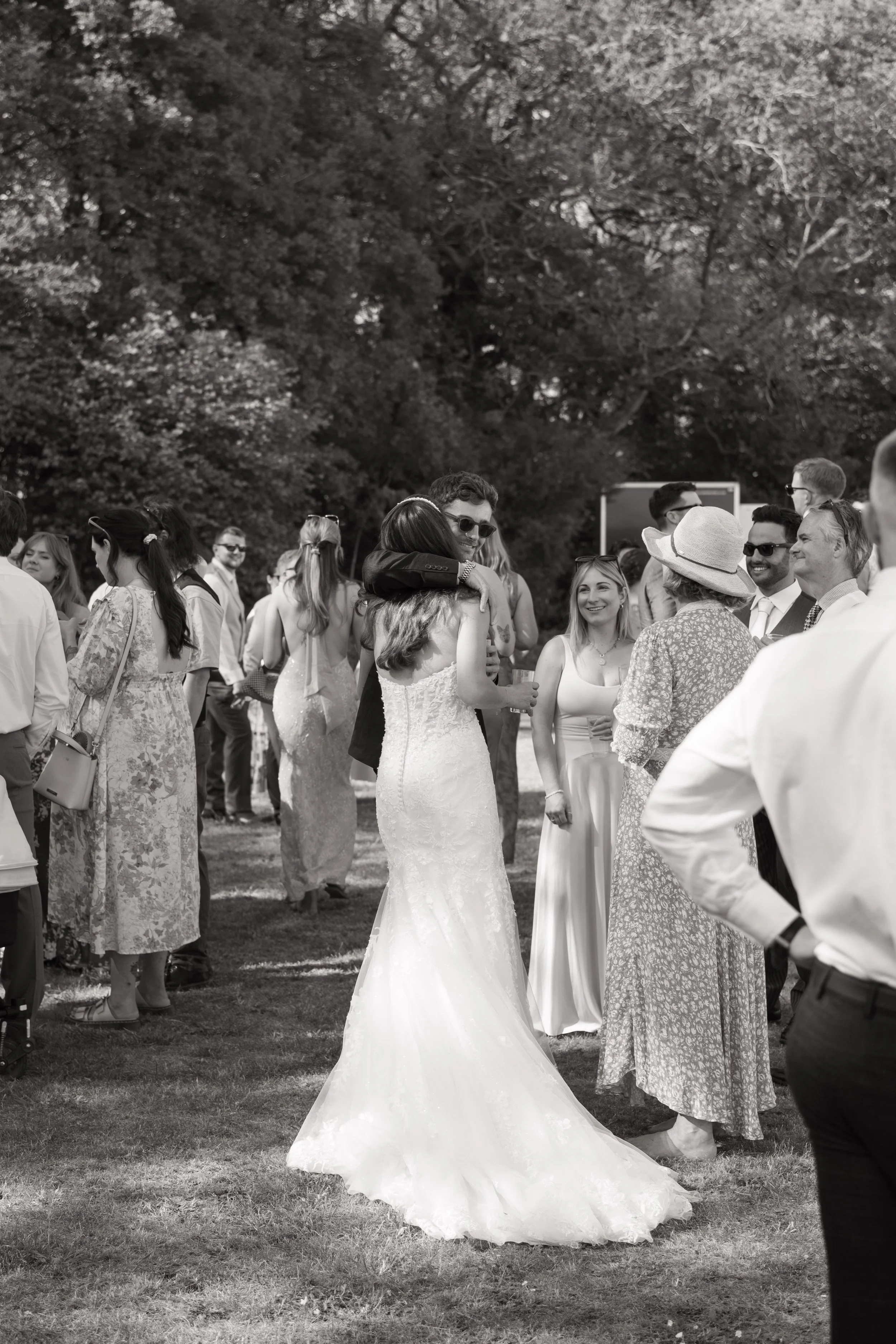 People gathered outdoors at a wedding event, with a bride in a white wedding gown and guests socializing and chatting.