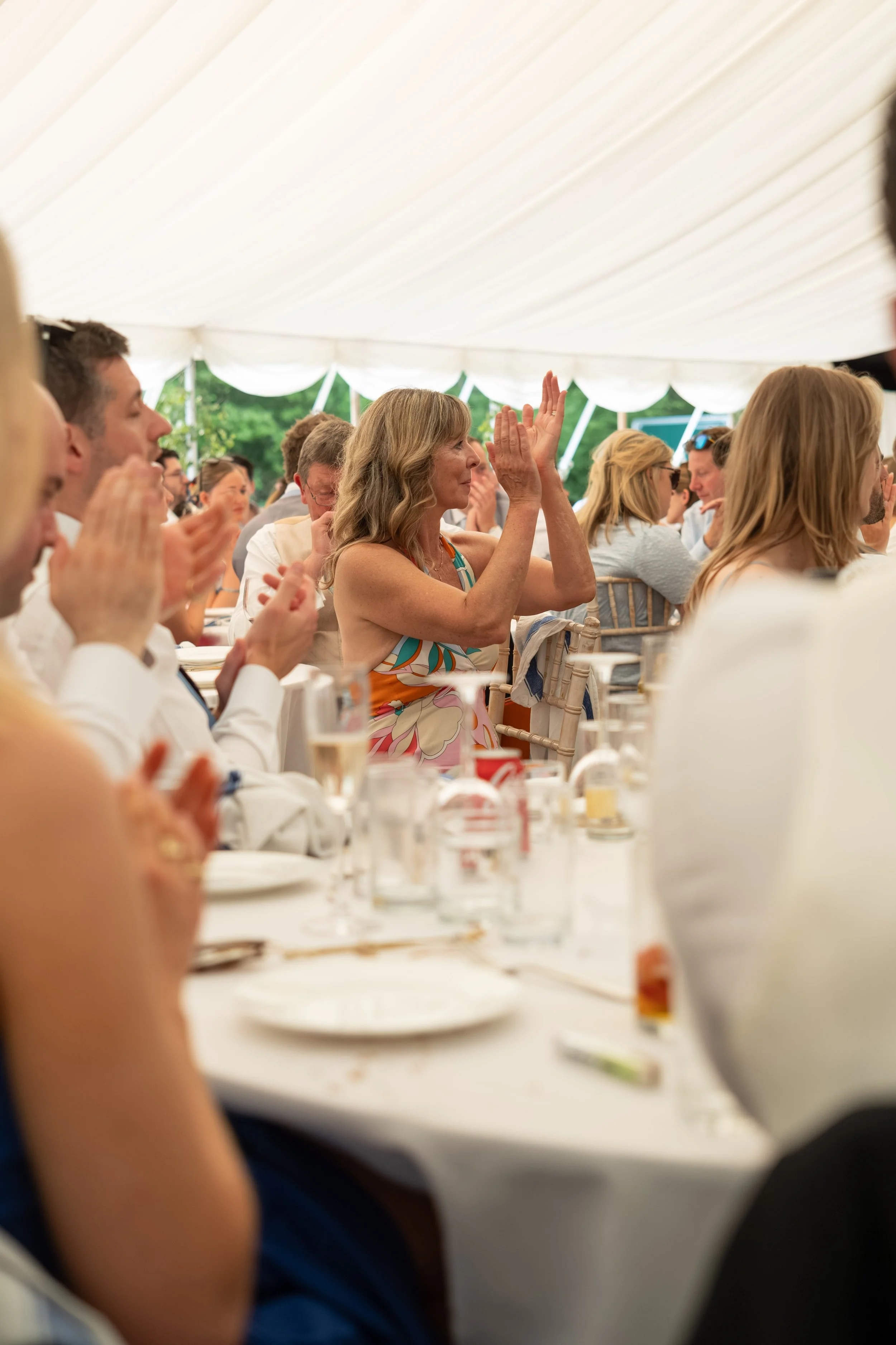 People seated at a table inside a white tent, participating in a formal event or gathering. Some are clapping, others are watching attentively. Drinks and table settings are visible.