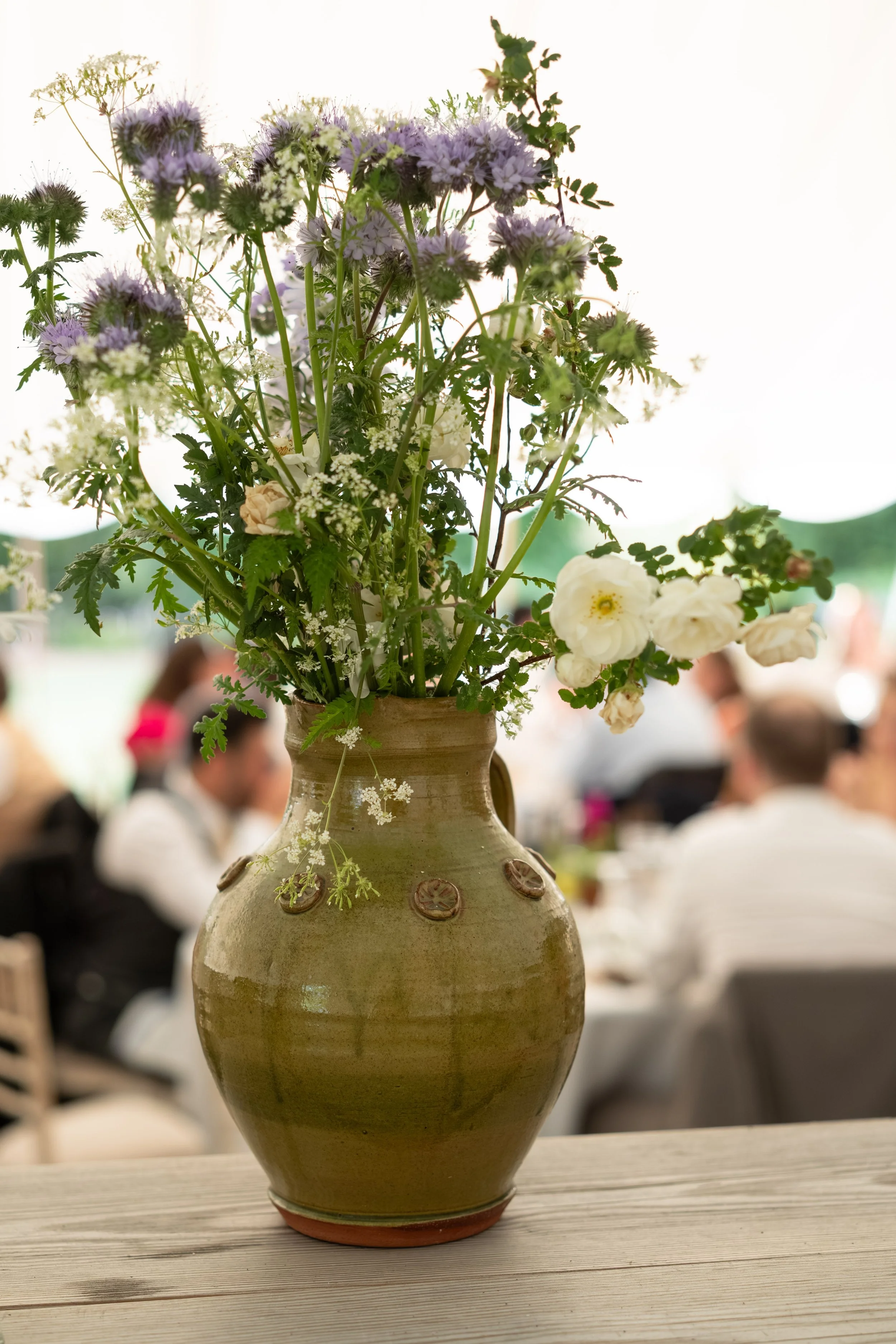 A large ceramic vase with a bunch of purple and white flowers, set on a wooden table, with a blurred background of a gathering of people at what appears to be a social event.