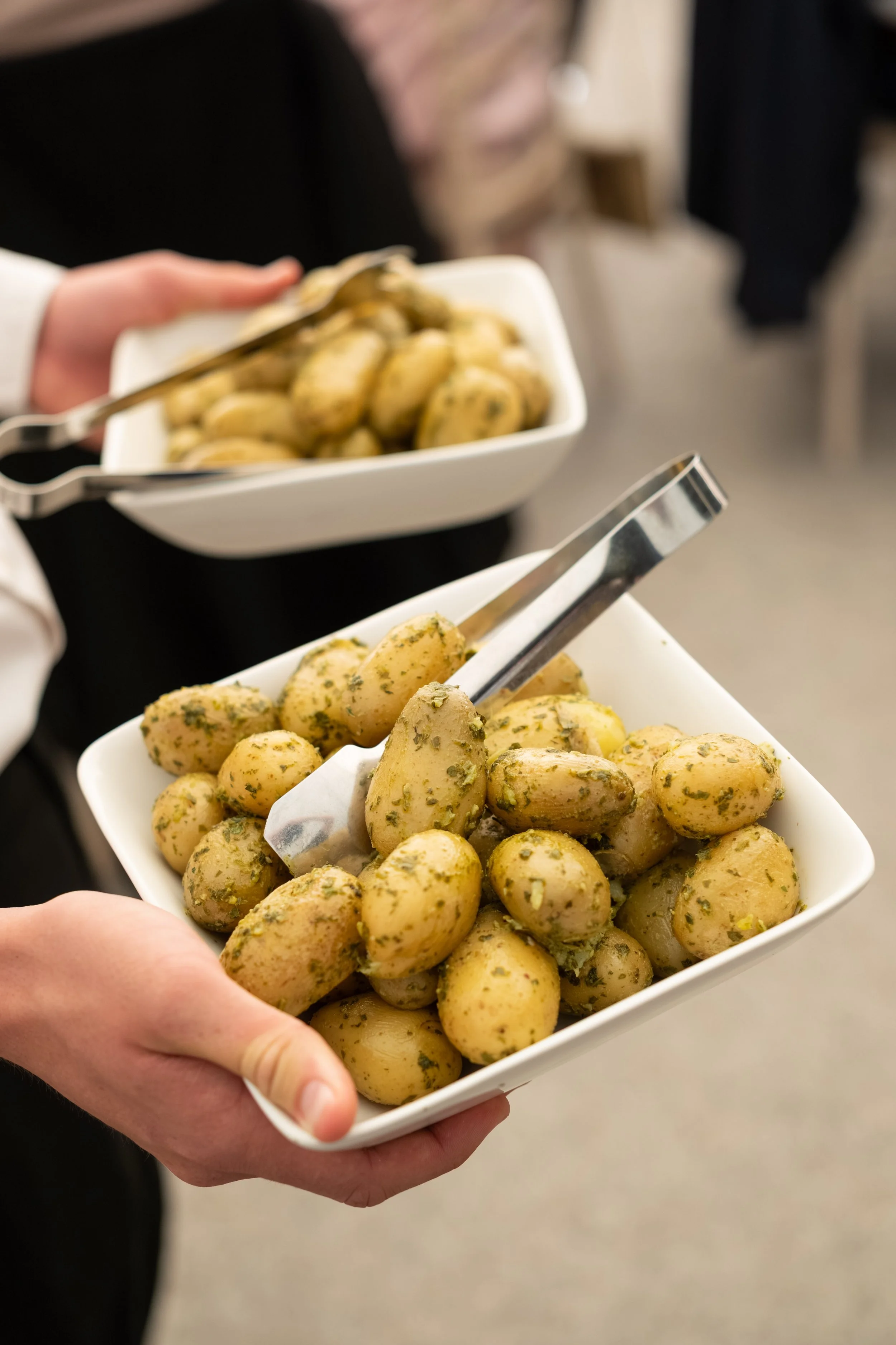Close-up of a hand holding a white rectangular bowl filled with small, herb-seasoned potatoes, with another similar bowl in the background.