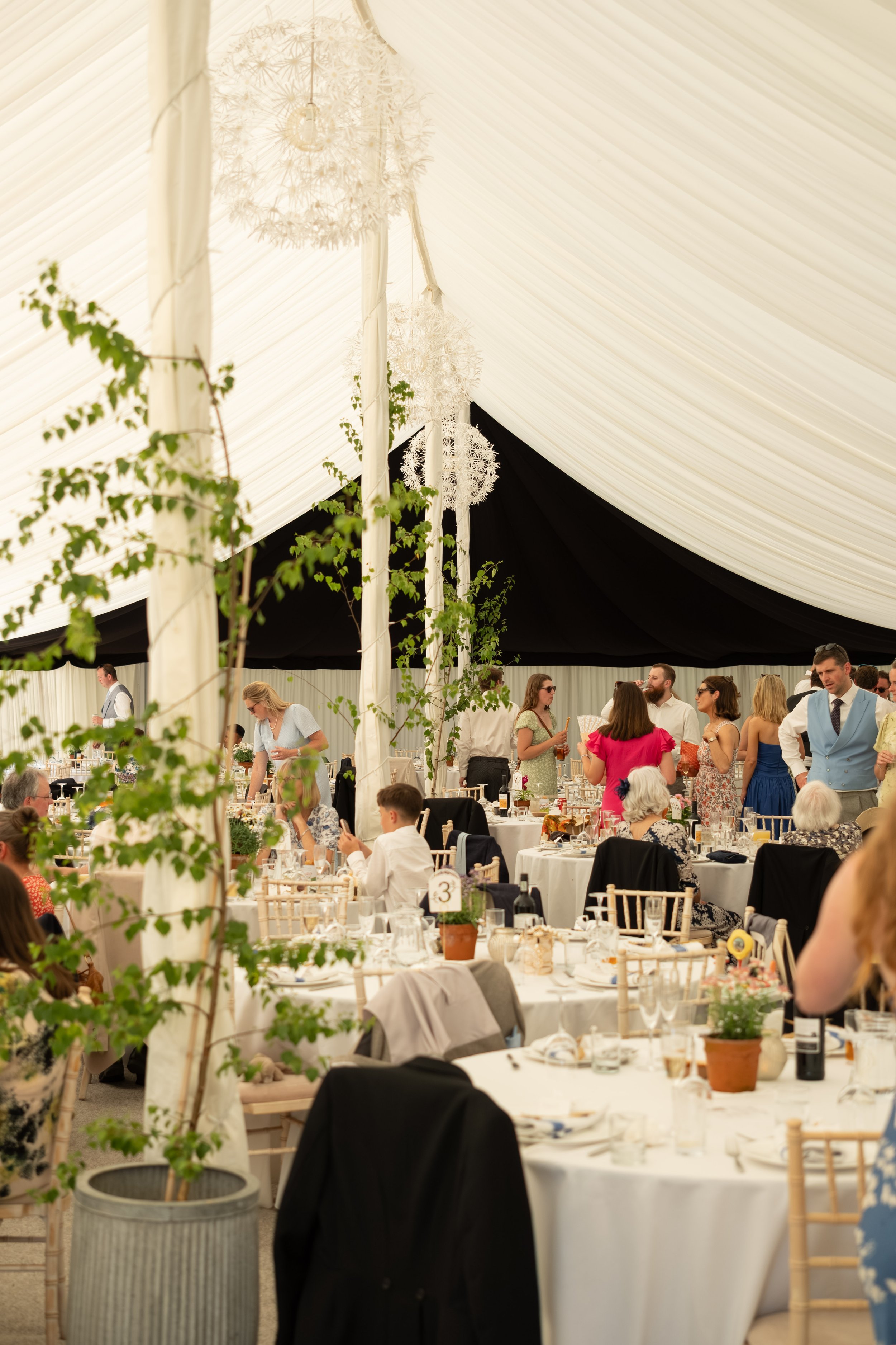 Guests standing and sitting at long tables with white tablecloths, dinnerware, and floral centerpieces inside a large tent decorated with white drapes and hanging white spherical chandeliers, attending a celebration or wedding reception.