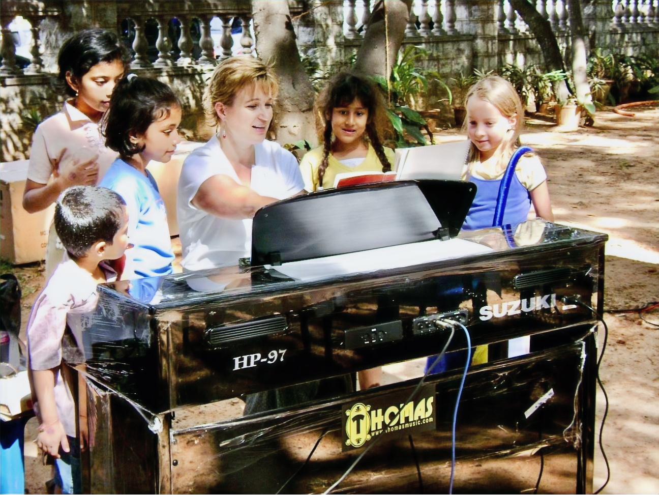 A group of young children and Andrea Humphrey gathered around a black Suzuki digital piano outdoors, with trees and a stone wall in the background.