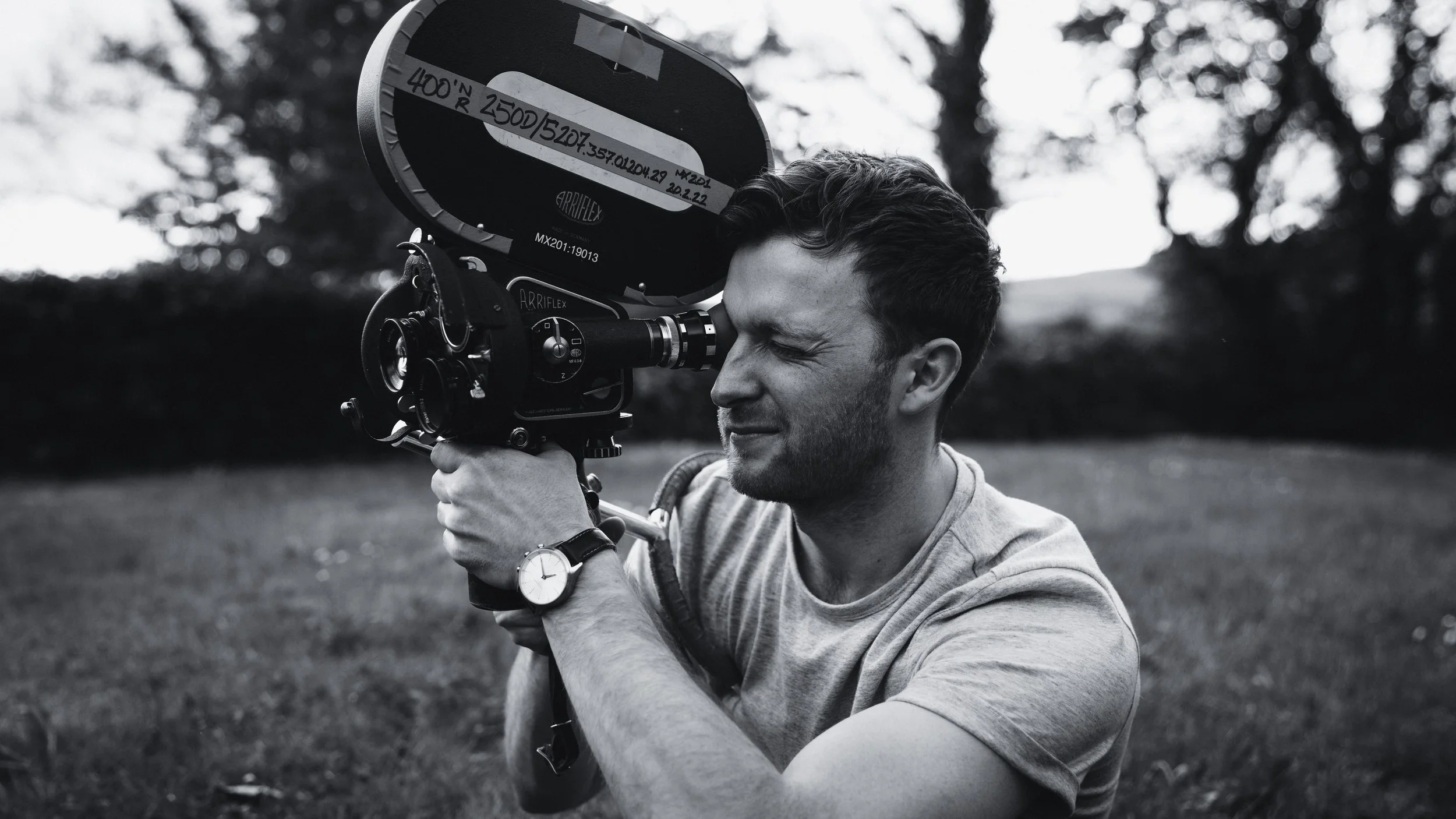 A man filming with a professional video camera outdoors, wearing a watch and a short-sleeved shirt, with trees in the background, in black and white.