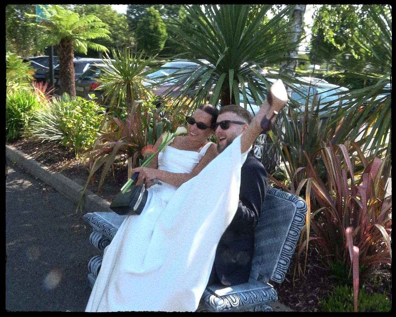 Two men are sitting on a park bench, laughing and smiling while one of them waves his arm. They are dressed in formal attire, wearing sunglasses, and are partially covered with a white cloth. They appear to be enjoying a sunny day in a landscaped area with plants, trees, and parked cars in the background.