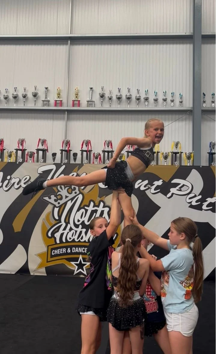 A cheerleading team performing a stunt in a gym with trophies on shelves in the background.
