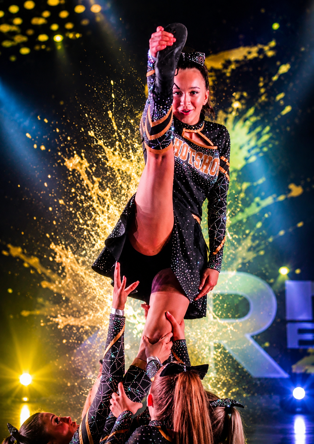 A cheerleading team performs a routine with a stunt, with a cheerleader lifted high in the air during a competition, with colorful lights and a sparkler effect in the background.