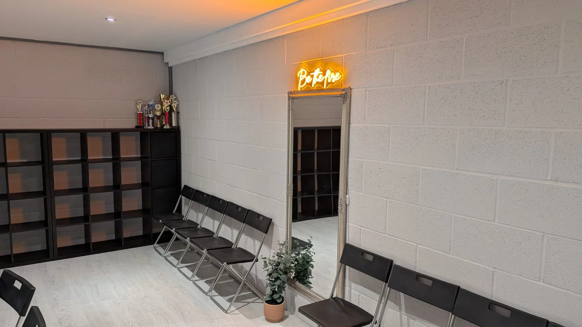 A waiting area with black chairs lined up against a white textured brick wall. There is a large mirror with an ornate frame and a neon sign that reads "Be Free" above it. On the left, there is a black open cubical shelf with trophies on top. There are potted plants on the floor near the chairs.