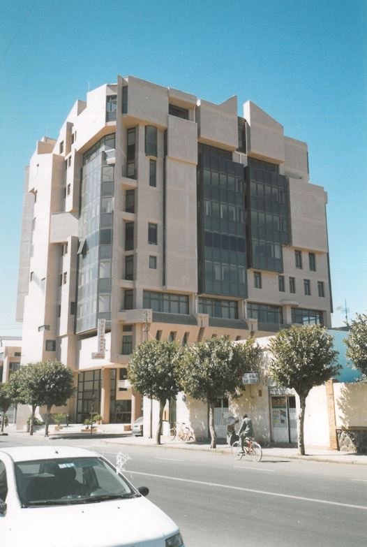 Modern multi-story building with a unique architectural design and large glass windows, located on a city street with trees and parked bicycles in front.