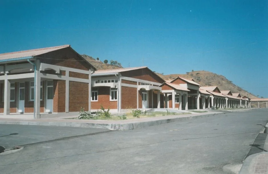 A row of identical single-story houses with brick walls and red tile roofs, situated on an empty street in a hilly area under a clear blue sky.