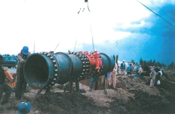 Group of workers installing a large black pipeline with red fittings in a dirt area, with several parked cars and trees in the background.