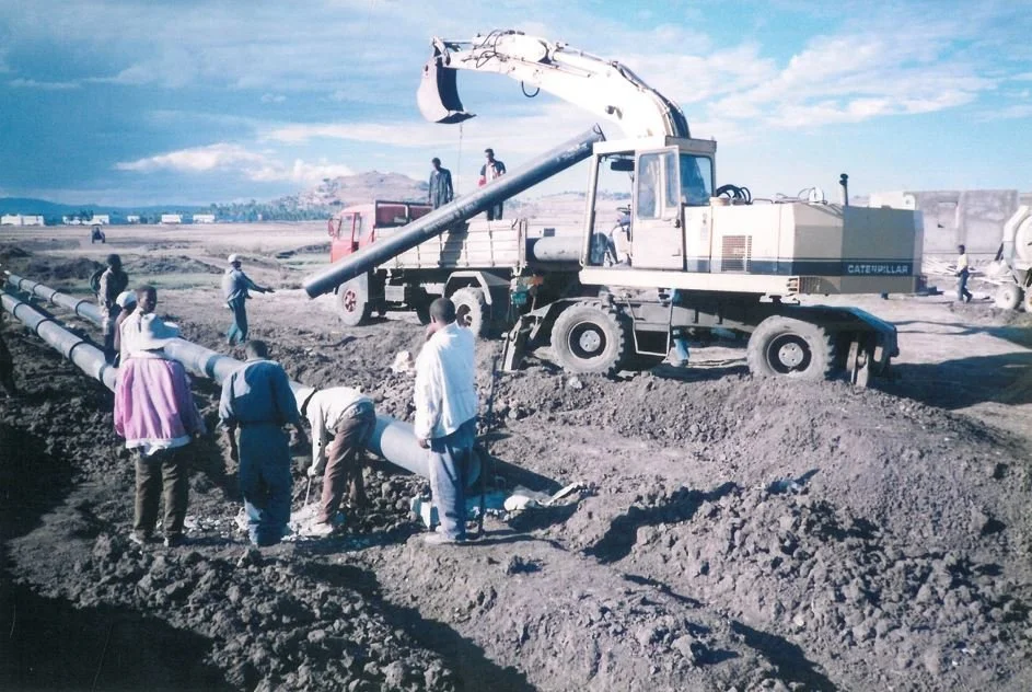 Construction workers installing a pipeline with a Caterpillar excavator on a dirt site under a blue sky.