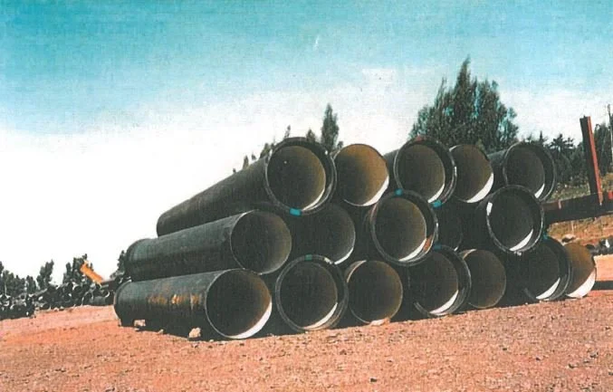 A stack of large black metal pipes lying on a dirt ground outdoors, with trees and a blue sky in the background.